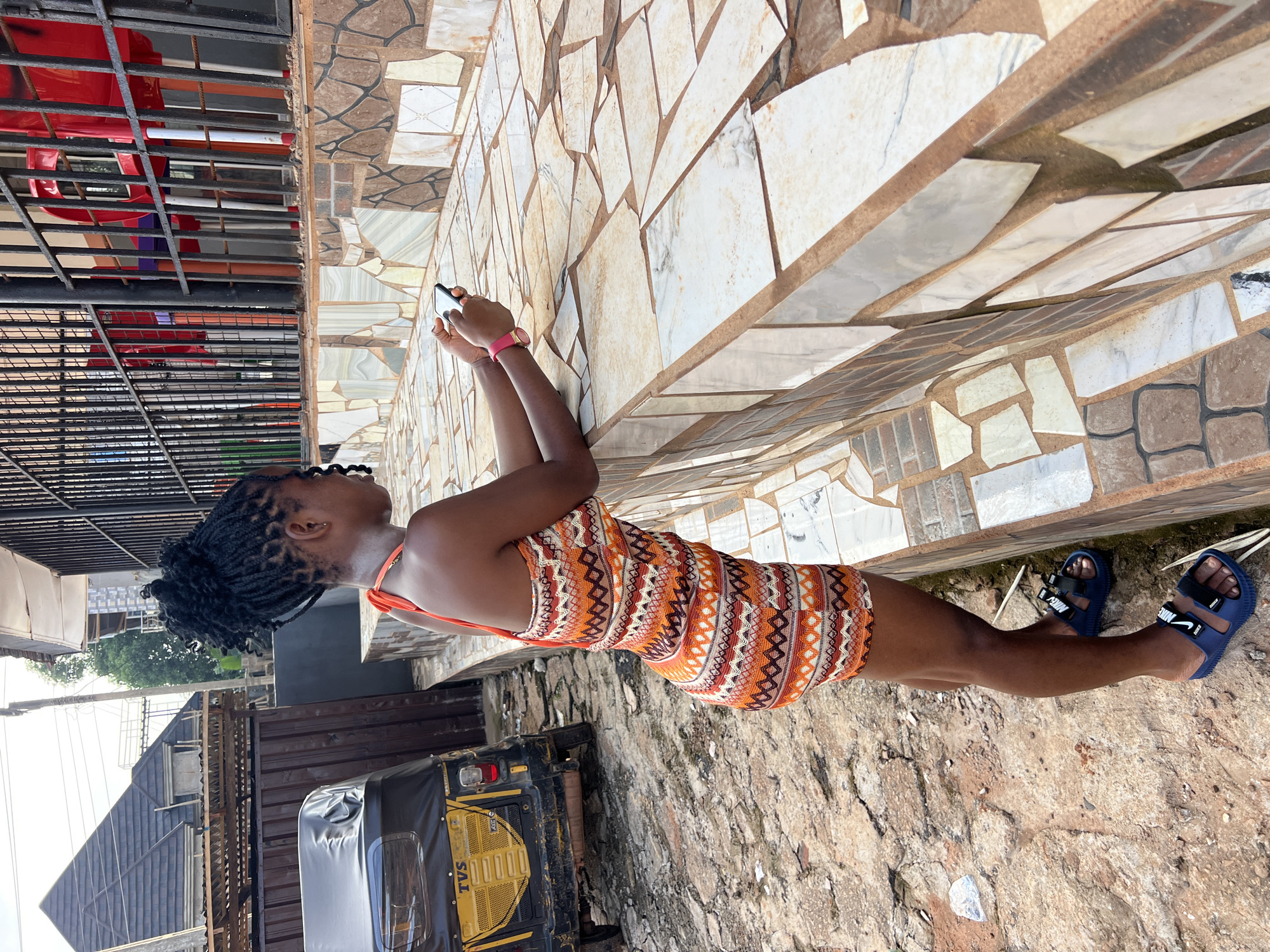 A young Black woman with dark skin and dreadlocks styled in a bun is standing outside, leaning on a tiled ledge, and looking at her phone. She is wearing an orange and white patterned sleeveless dress and blue slide sandals. In the background, a yellow and black auto-rickshaw is parked on a dirt and stone path. Behind the woman, a black metal fence encloses a seating area with red chairs visible. The environment appears to be a street or market area in Nkpor, Nigeria, indicated by the architecture, vehicles, and general streetscape. The lighting suggests it is daytime, possibly late afternoon due to the angle of the sun.