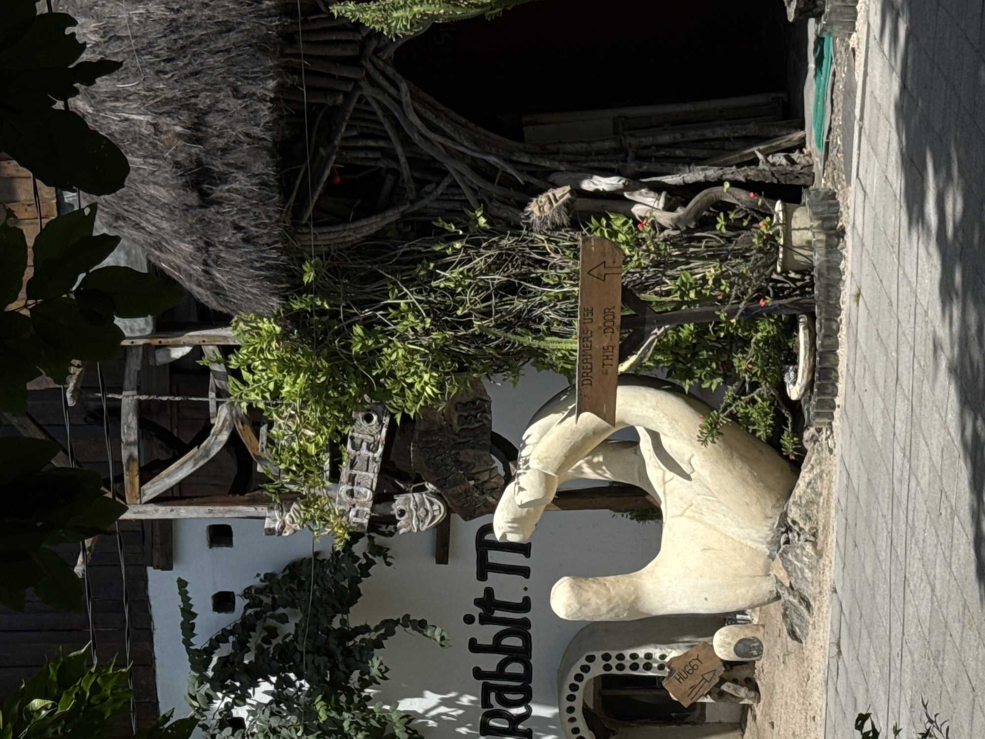 Here is a description of the image:
The image shows the exterior of "Rabbit Town" in an area that appears to be Unknown, Indonesia. The storefront is white with "Rabbit Town" written vertically in black. A large, white hand sculpture extends from the wall. A sign reading "Dreamers use this door" with an arrow is visible. Another sign reads "Huggy" with an arrow. Plants are growing on the facade of the building. The sidewalk in front is tiled and has shadows. The upper portion of the building features a rustic design with wood and thatched elements.
