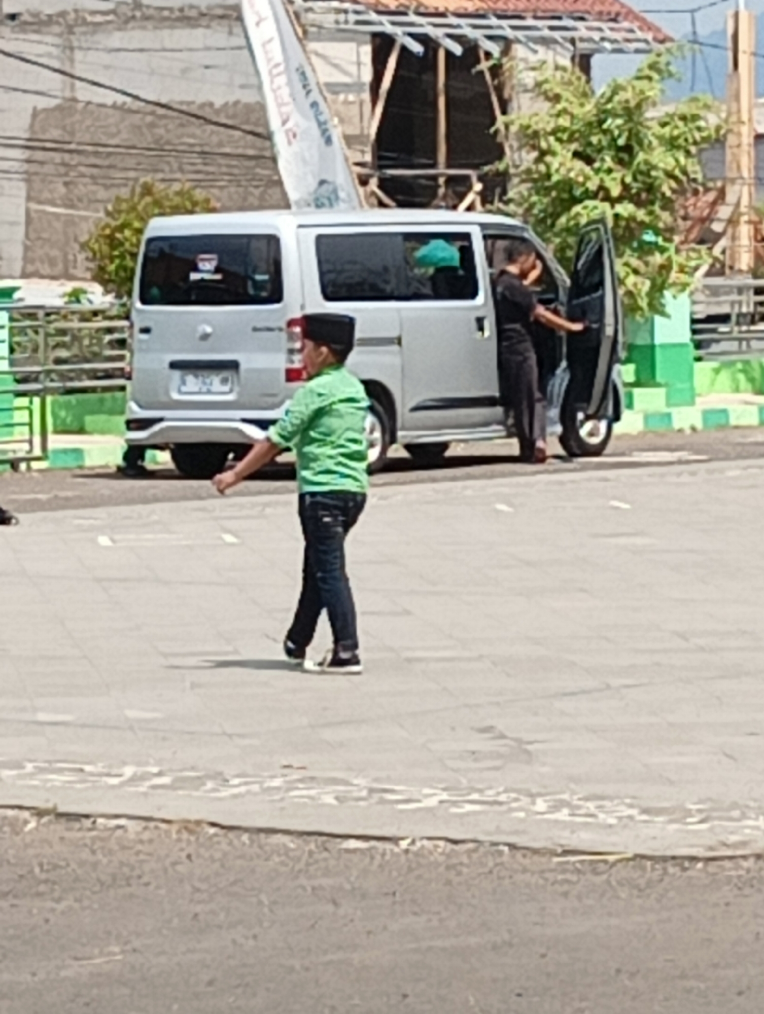 A young boy wearing a green shirt, black pants, and a black cap walks in an open, paved area. To his right, a silver van is parked with its side door open, and two individuals, one in a black shirt and black pants, and another in a black shirt and dark pants, are standing by it. The environment suggests an outdoor setting in Kuningan, Indonesia, with a building and foliage visible in the background. The paving stones appear weathered, and there are some low green barriers in the background. The lighting indicates it is daytime.