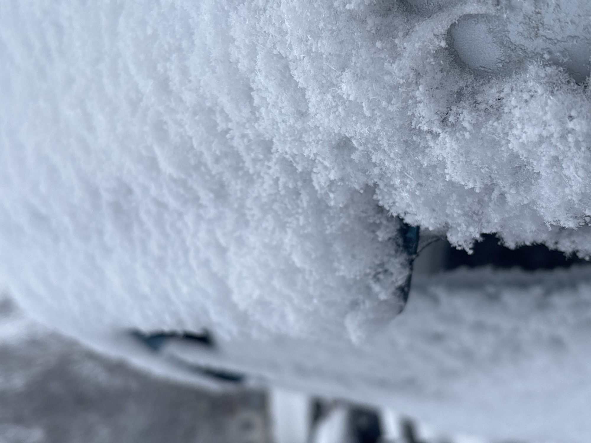 A close-up shot displays a surface heavily covered in fresh snow. The snow appears crystalline and textured, obscuring the object it blankets. Due to the extreme close-up and lack of discernible landmarks, it is impossible to confirm the location as Izhevsk, Russia. The scene primarily showcases the snow itself and its texture.