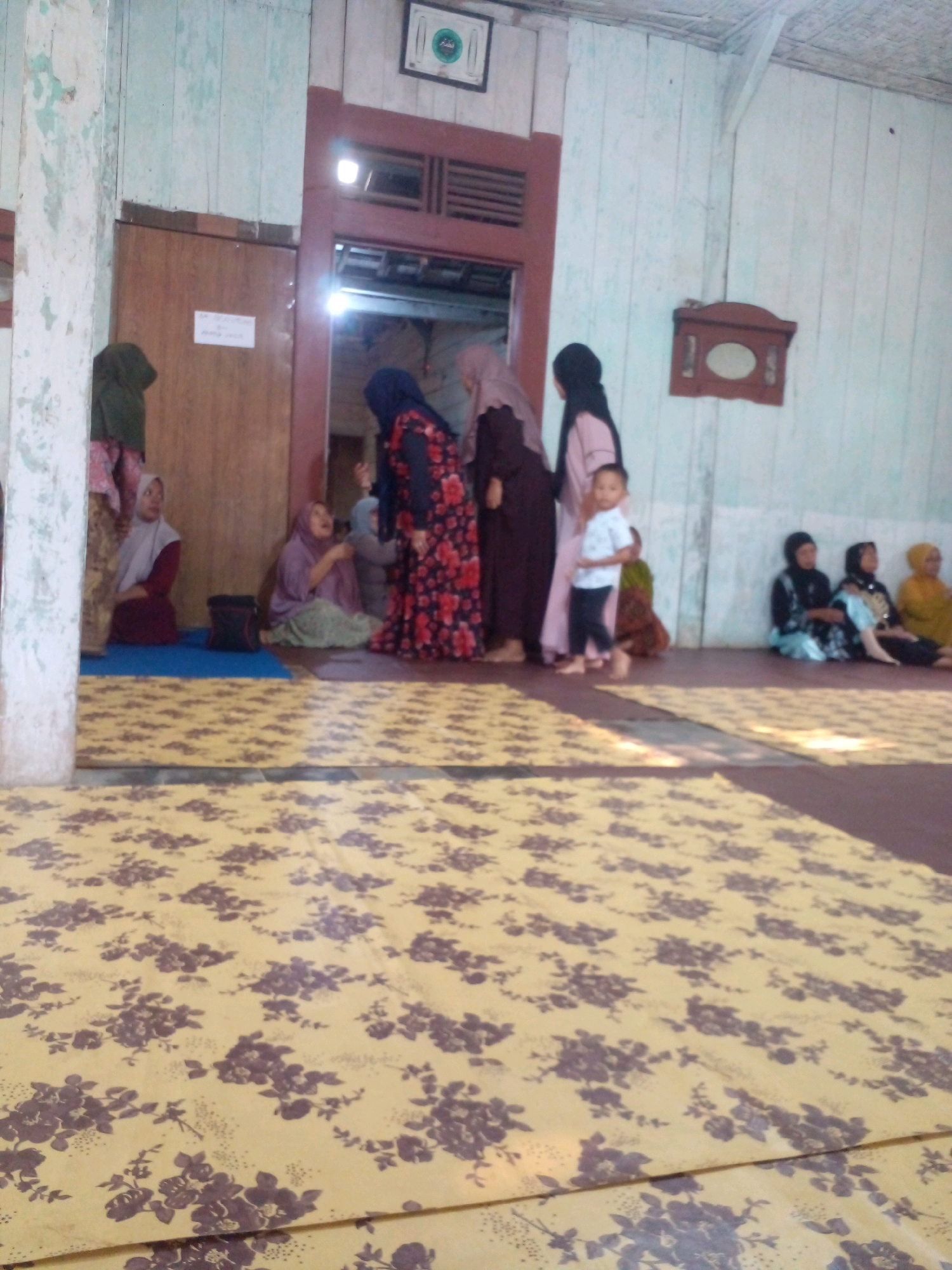 The image depicts an indoor gathering, possibly in a community hall or a large room within a home, located in Pungangan, Indonesia. The environment appears somewhat rustic, with light-colored wooden walls and a doorway leading to another area. The floor is covered with a large, yellow mat adorned with a floral pattern of dark purple flowers.
Several individuals are present, mostly women and children, dressed in modest attire typical of Indonesian cultural norms, with many wearing headscarves. On the left side of the image, two women are seated on the floor, one wearing a maroon hijab and the other a purple hijab. Behind them, a dark bag is placed on the floor. In the center of the room, a woman in a vibrant red and black floral dress is standing, interacting with others. A young boy in a white shirt and dark pants is also visible, appearing to be walking or standing. Further to the right, a group of women are seated together. One woman wears a black hijab, another a blue headscarf, and a third a yellow hijab. The overall scene suggests a social or religious gathering, possibly a family event or community meeting. The lighting is subdued, with some ambient light entering from an open doorway and possibly from overhead sources.