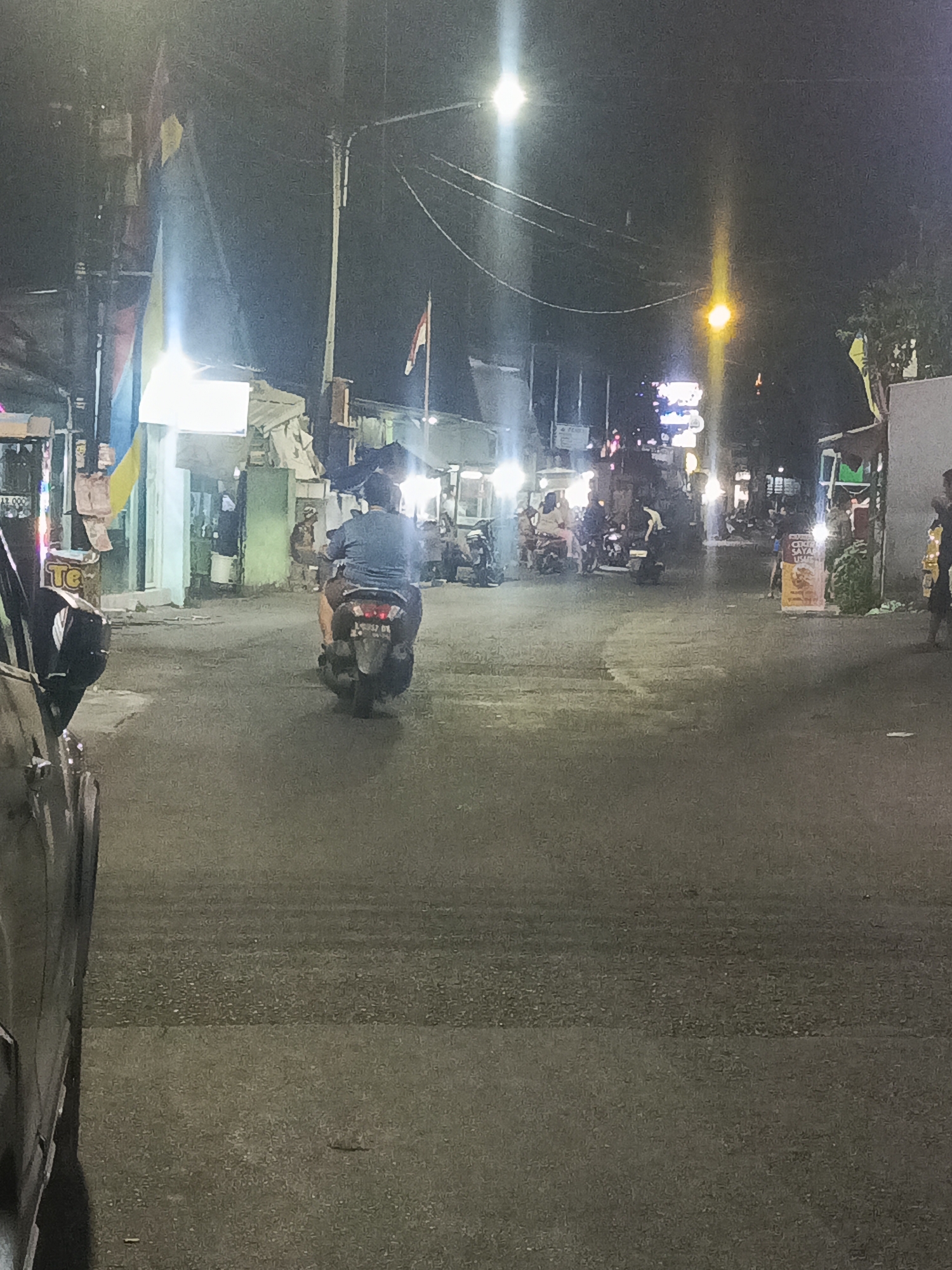 The image depicts a nighttime street scene in Surabaya, Indonesia, likely in an urban or semi-urban area given the presence of shops and streetlights. A person is riding a motorcycle down the asphalt road. The motorcycle's taillight is illuminated. On the left side of the road, a dark-colored vehicle, possibly a car, is partially visible. In the background, several small shops or stalls line the street, with some illuminated by bright lights. Other motorcycles and people are visible in the background, suggesting moderate activity. Various wires and streetlights crisscross overhead. Flags are visible on poles near the shops on the left. The overall atmosphere is one of a typical busy street at night in a Southeast Asian city.