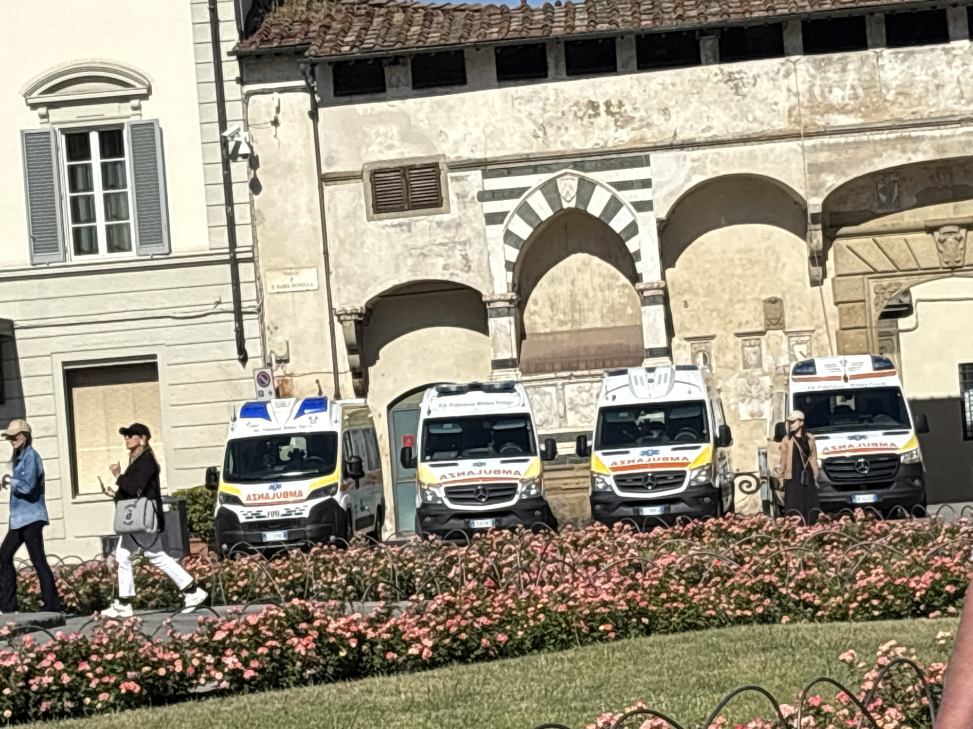 The image depicts a scene in Florence, Italy, with four ambulances parked in front of a historic building with arches. The ambulances are white with yellow and blue accents and bear the logo "ASHA JURMA". Two pedestrians are visible: one woman in a denim jacket is walking left and another wearing a black jacket and white pants is walking to the right. A bed of pink flowers lines the foreground, and a wrought iron fence separates the flowers from the viewer. The building in the background features a mix of architectural styles, including a window with gray shutters on the left and arches decorated with black and white stripes in the center. The facade of the building appears aged and weathered.