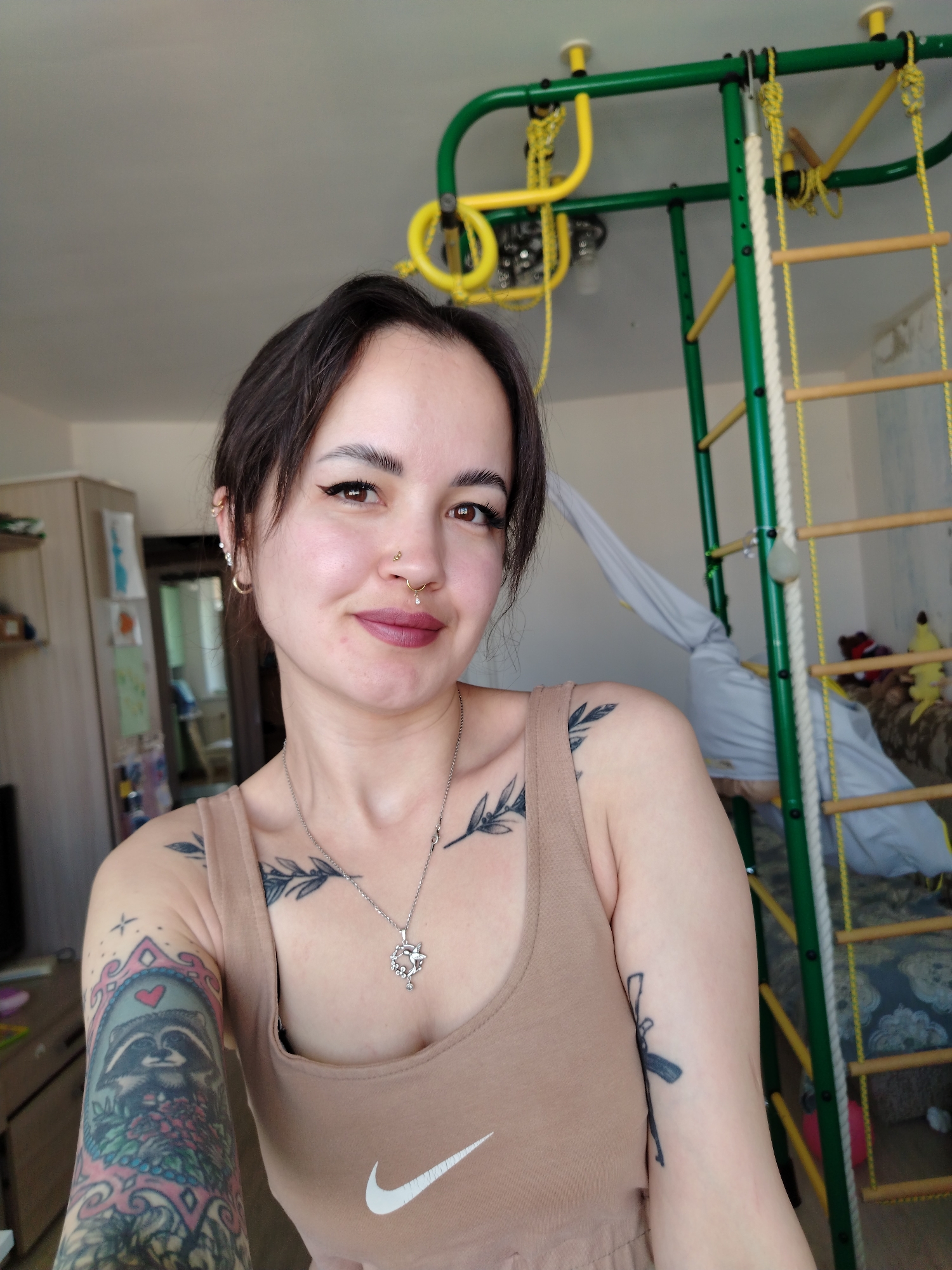 A fair-skinned woman with dark hair pulled back is centered in the frame, appearing to take a selfie. She has multiple piercings in her left ear and a nose ring. She wears a tan tank top with a white Nike logo. Tattoos are visible on both arms and chest. The background features a child's indoor play structure with green, yellow, and white elements. A cabinet is visible to the left, and a curtained area and other furniture are partially visible behind the play structure. The location appears to be an apartment in Noviy, Russia.