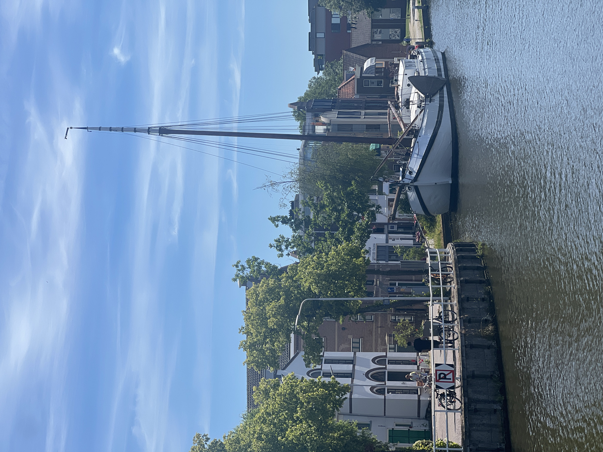 The image depicts a serene canal scene in Delft, Netherlands. A white sailboat with a tall mast and rigging is moored alongside a canal. The boat is long and slender, with a white hull and dark trim. On the left side of the canal, a waterfront promenade is lined with buildings, trees, and bicycles. The buildings are a mix of architectural styles, with white facades and arched windows. Several people are visible on the promenade, some walking and some standing near the bicycles. The sky is blue with scattered clouds, and the water of the canal is dark and reflective. The scene is illuminated by bright sunlight, creating a peaceful and idyllic atmosphere.