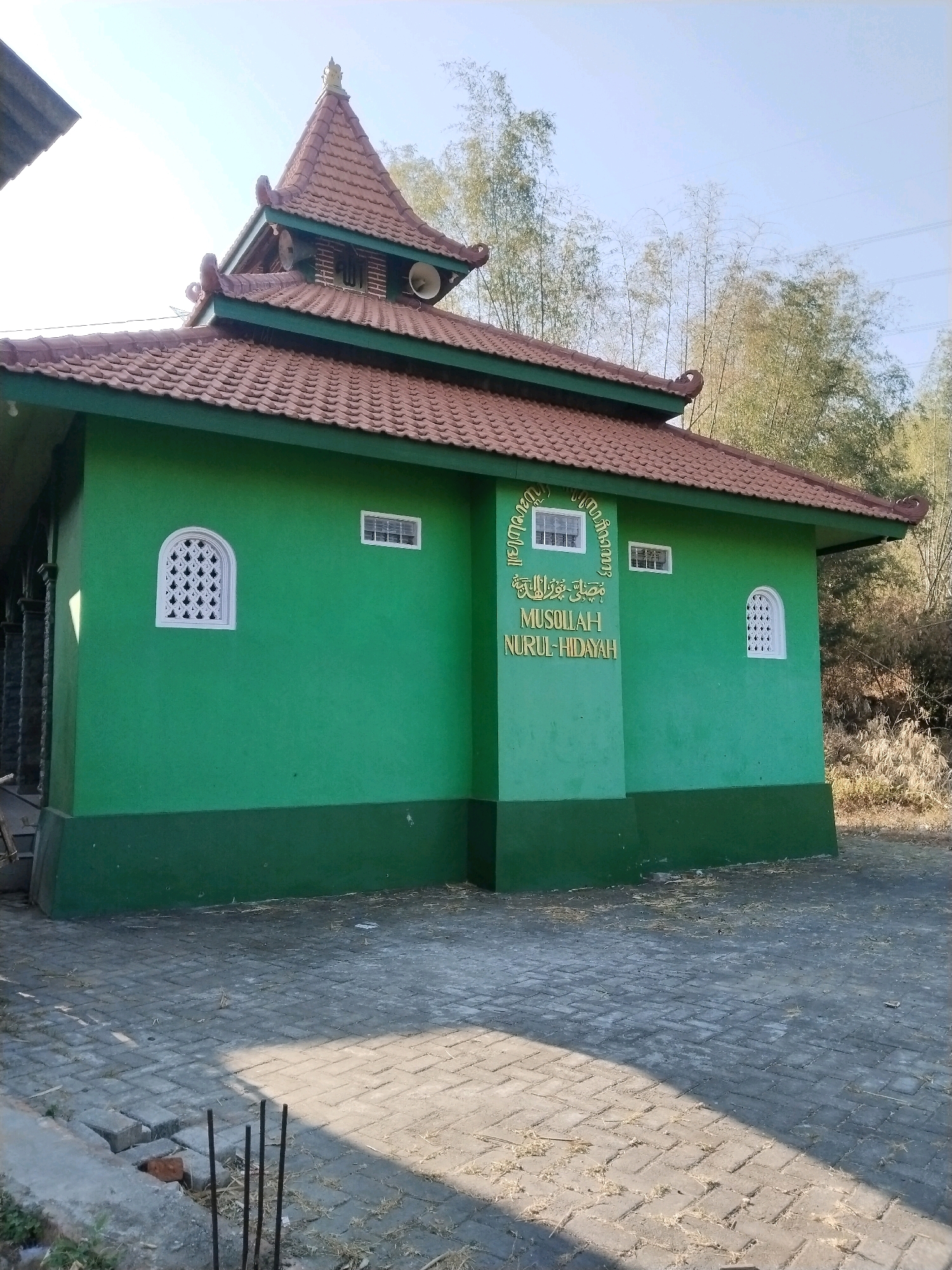 A vibrant green, single-story mosque with a brown tiled roof stands under a clear blue sky. The mosque features several white windows, one with an ornate, decorative design. Arabic calligraphy and the text "MUSOLLAH NURUL-HIDAYAH" are inscribed on the front of the building. A speaker is visible on the roof. The mosque is surrounded by a paved area with scattered dry leaves. In the background, tall bamboo plants are visible. The image appears to be taken in a rural or suburban setting, possibly in Kuripansari, Indonesia, given the context provided. There are no people in the image.