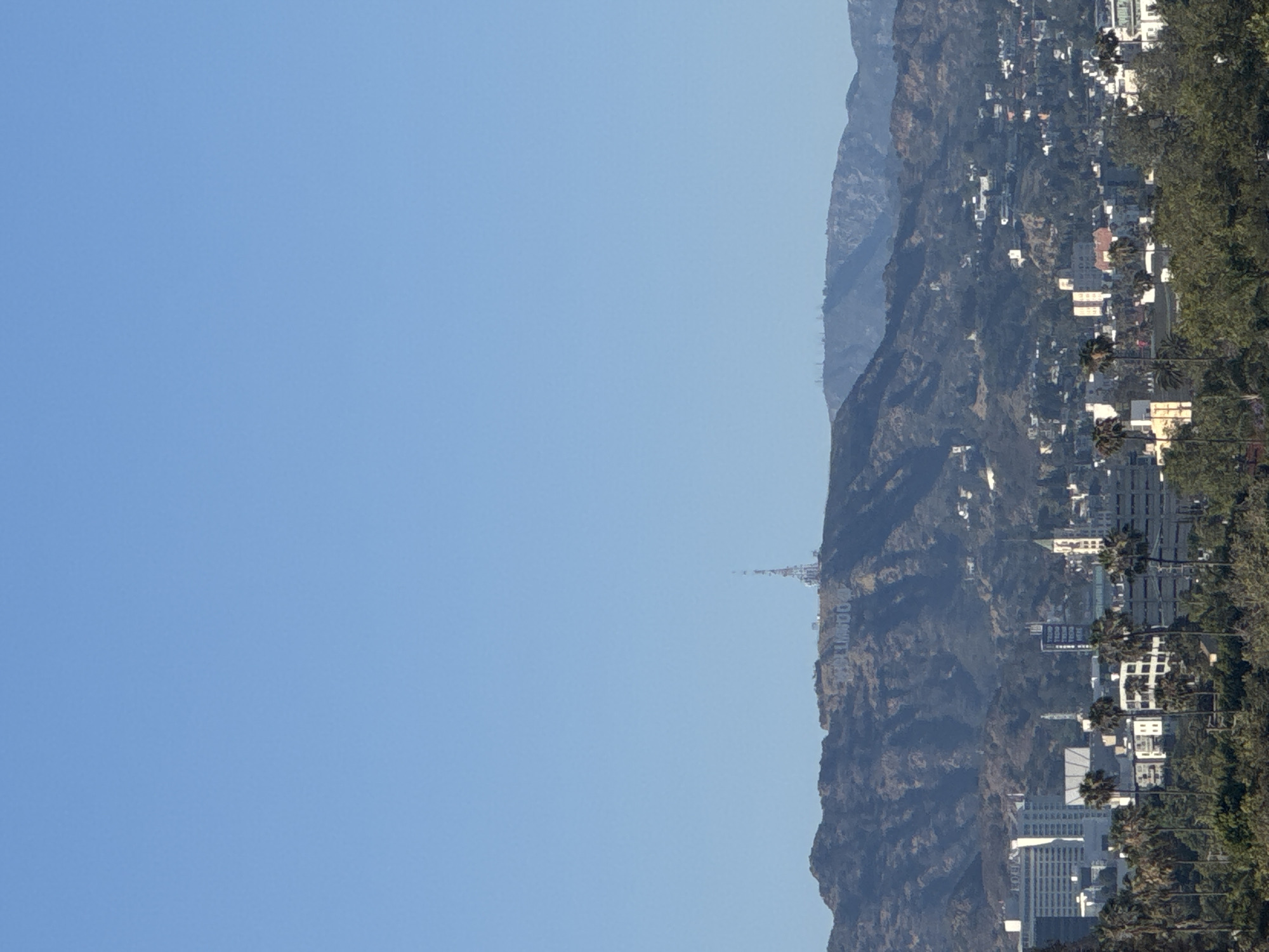 This image shows a daytime view of Los Angeles, California. The Hollywood Hills are visible, populated with residential and commercial buildings. The Hollywood sign can be seen on the hillside. The sky is clear and blue.