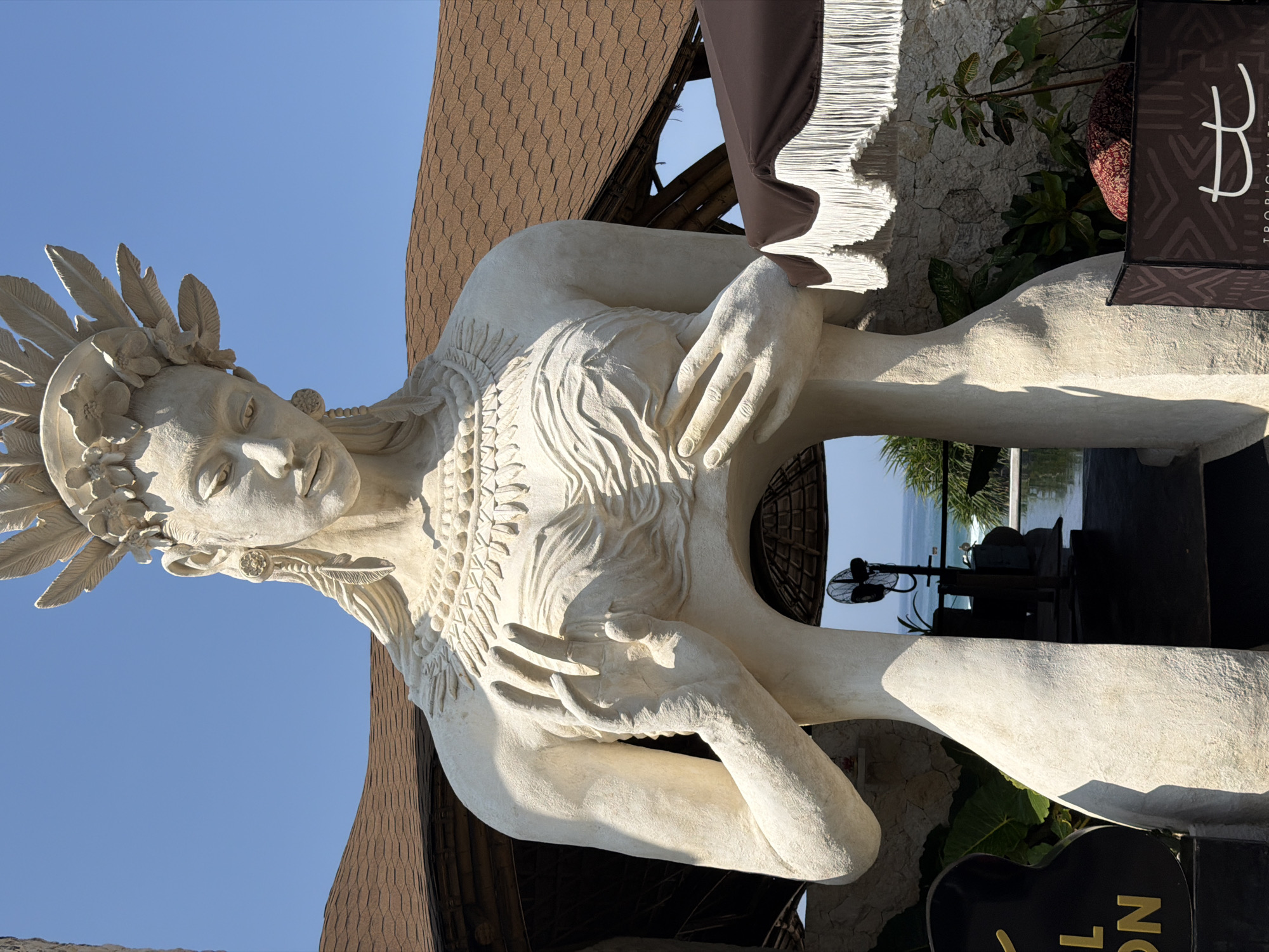 A large, light-colored, stylized female statue stands prominently outdoors under a clear blue sky in Ungasan, Indonesia. The statue features an elaborate headdress composed of feathers and floral elements, a serene facial expression looking downwards, and detailed patterns resembling jewelry on its neck and chest. The mid-section of the statue's torso is hollow, forming an archway. Through this opening, a distant blue ocean with white waves and a horizon line is visible, along with a black pedestal fan, green foliage, and a dark reflective surface, possibly a pool. The statue's left hand is raised with splayed fingers in a specific gesture, while its right hand rests over the breast area. Behind the statue, a building with a brown shingled roof and textured stone walls is present. To the right of the statue, a partial view of a brown umbrella with white fringe and a dark stand featuring tribal patterns and the letters "tt" (suggesting "Tropical T...") is observed. To the left, a black sign with partially visible yellow lettering is visible.