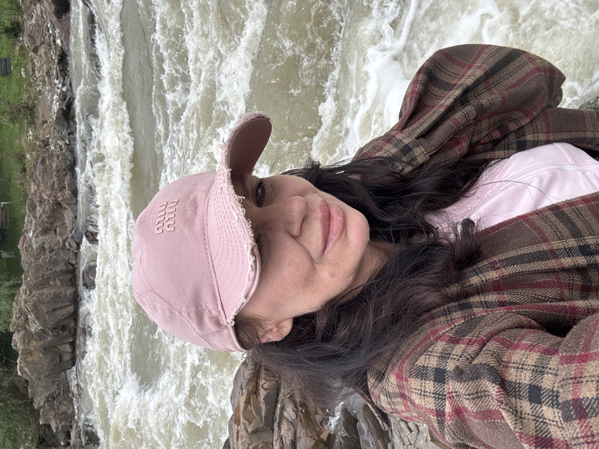 A woman poses for a selfie with a rushing river in the background. She is wearing a pink baseball cap, a pink shirt, and a brown plaid jacket. The river is turbulent and appears to be flowing over rocks. The location is likely near a body of water in Каменномостский, Russia.