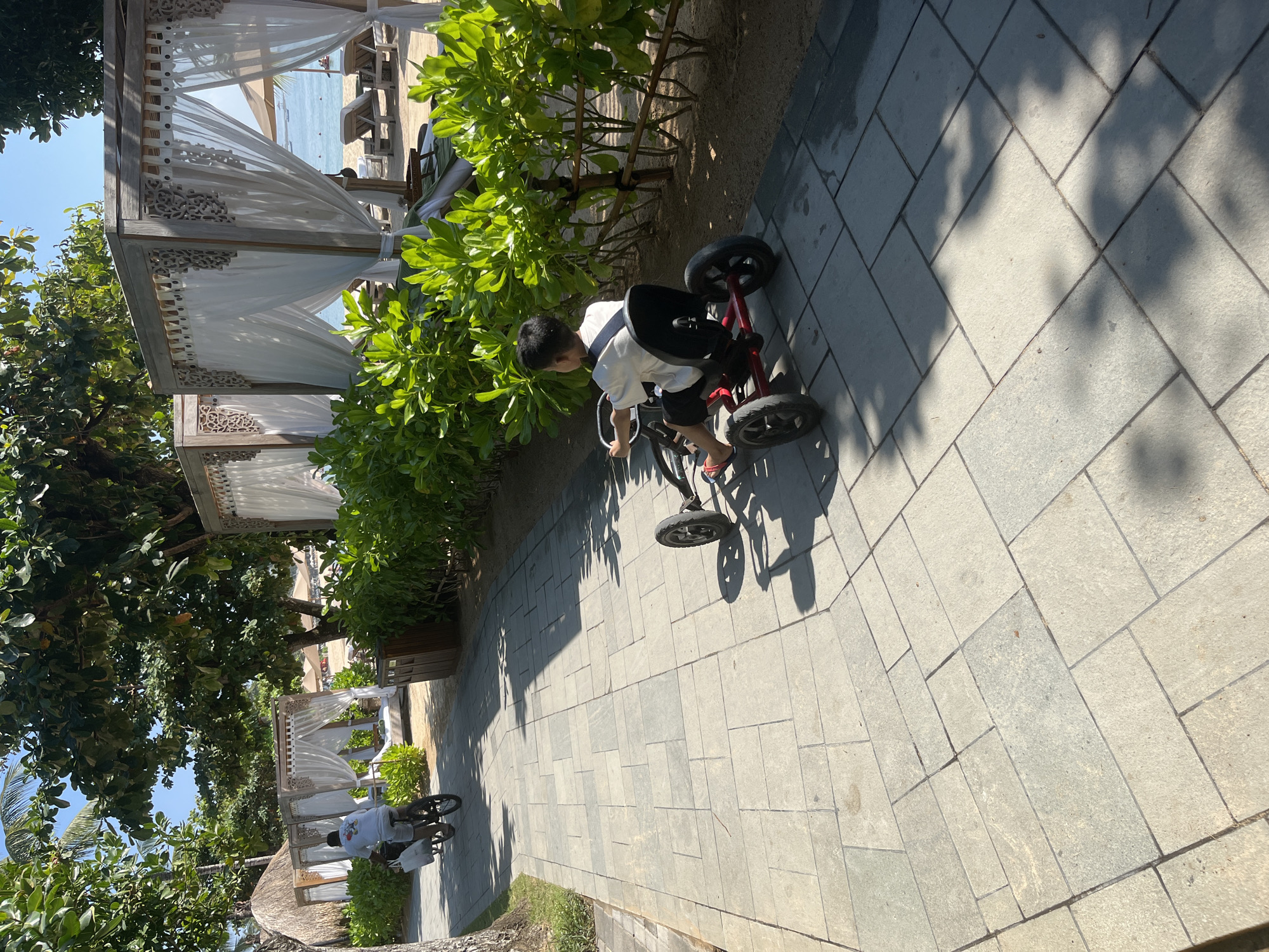 A young boy rides a red pedal go-kart on a paved walkway at the beach resort in Kuta Selatan, Indonesia. He is wearing a white t-shirt and black shorts, and a black strap is around his chest. His feet are on the pedals. In the background, a person on a bicycle is also on the walkway. The walkway is bordered by lush green bushes. Beyond the bushes, beach cabanas with white drapes are visible, as well as beach chairs on the sand and the ocean. The sky is clear blue. The sun is casting shadows on the walkway.