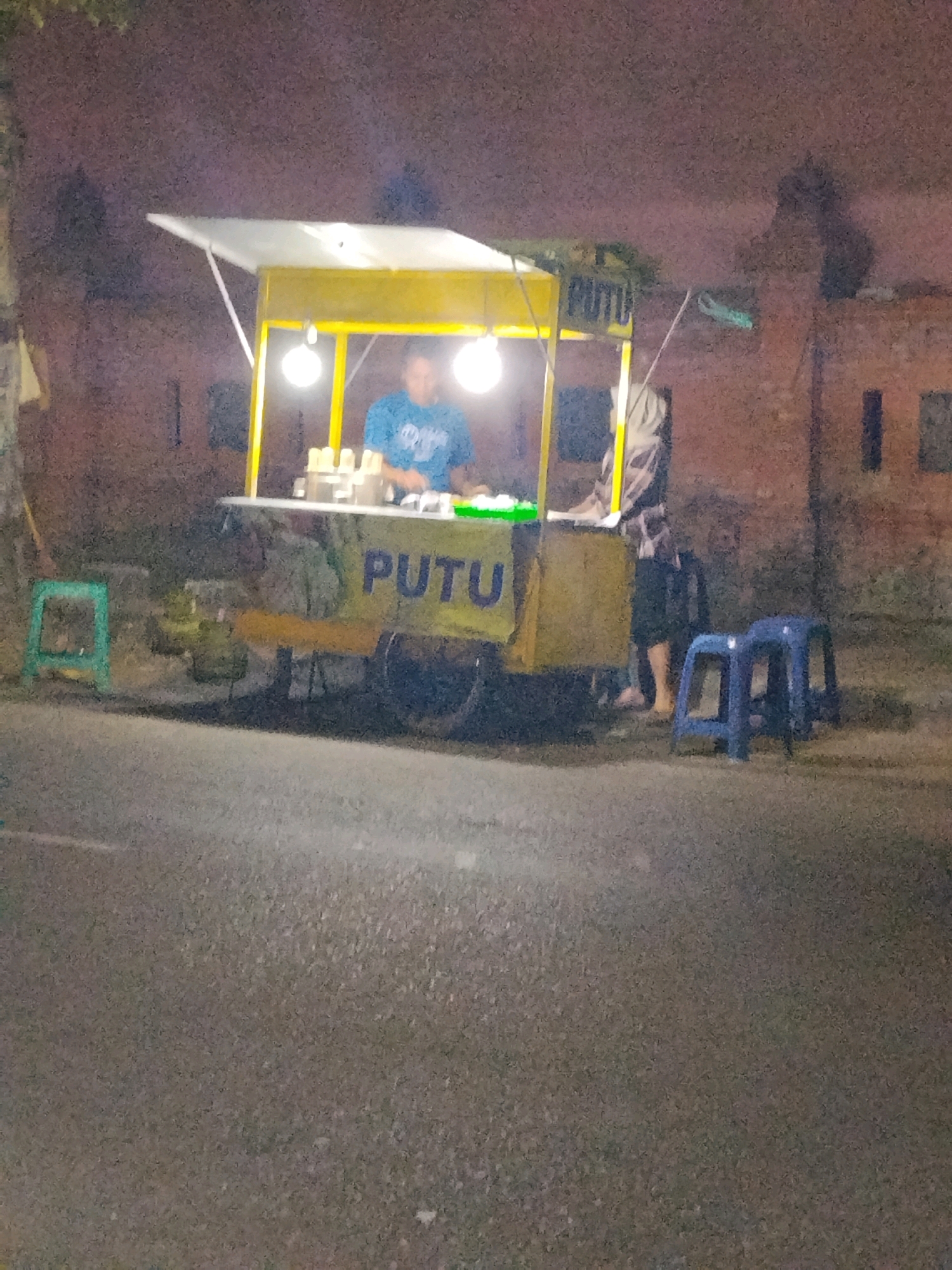 A food cart with a yellow awning is parked on the side of a street at night in Mojokerto, Indonesia. A person wearing a blue shirt stands behind the cart, preparing food. The cart has a sign that reads "PUTU." Two blue plastic stools are placed in front of the cart, and a green stool is to the left of the cart. The street is made of asphalt, and brick buildings are visible in the background. The scene is illuminated by streetlights and the lights from the food cart.