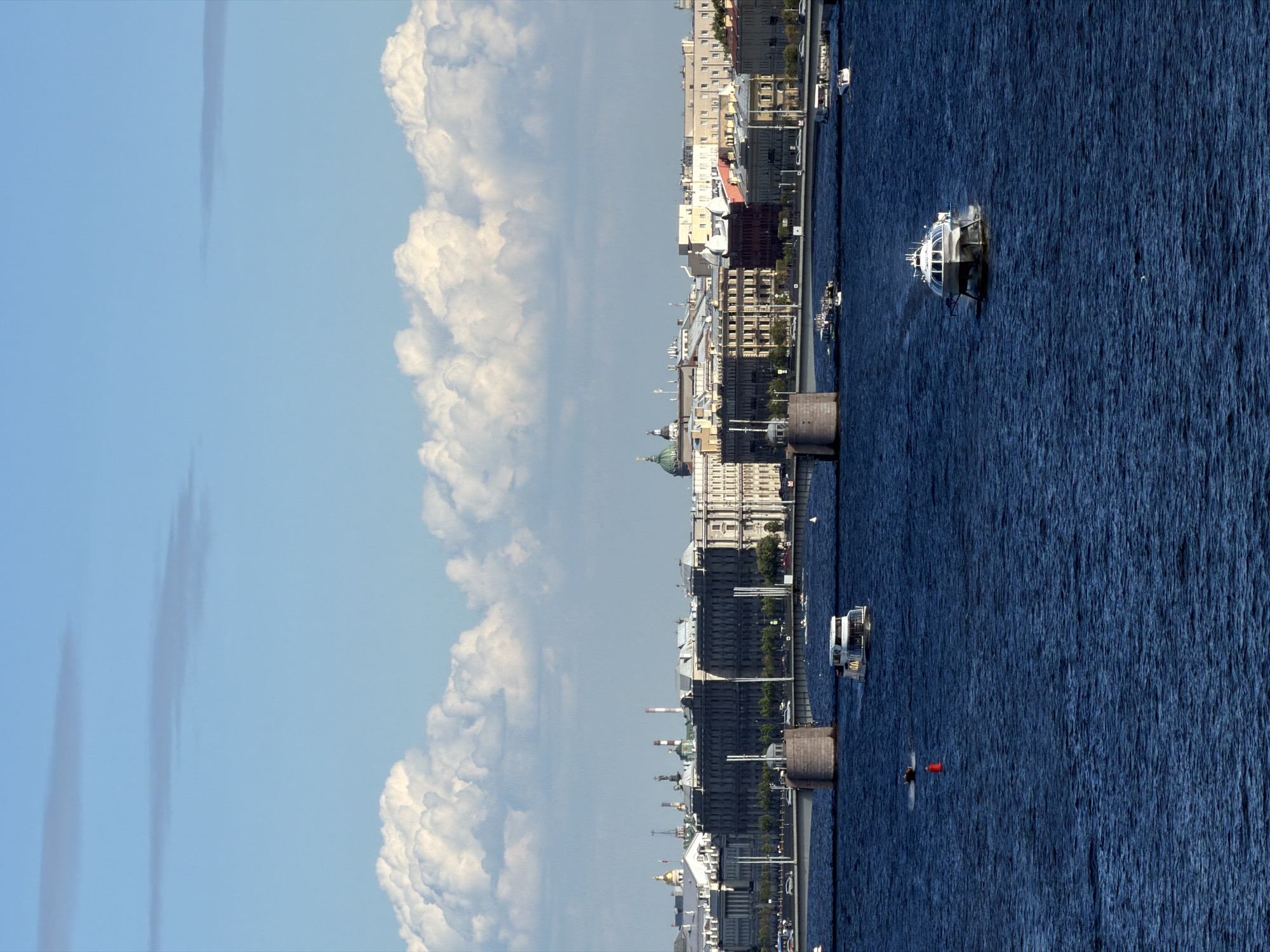 The image captures a panoramic view of the Neva River in Saint Petersburg, Russia, under a bright, partly cloudy sky. In the foreground, the deep blue water is choppy, with several boats navigating its surface. A prominent white hydrofoil, resembling a sleek, modern vessel with a glass-like cabin, is moving towards the viewer from the right. Further left, a smaller white boat is sailing, and a red buoy is visible in the water between the boats.
In the middle ground, a wide bridge spans the river, with stone piers supporting its structure. Cars and pedestrians can be seen on the bridge, indicating a busy thoroughfare. Behind the bridge, the St. Petersburg skyline unfolds, characterized by a collection of classical and historical buildings. Notable structures include buildings with ornate facades, domes, and spires, characteristic of the city's architecture. The historical grandeur of the cityscape is accentuated by the presence of what appears to be the Rostral Columns in the distance, a landmark in St. Petersburg.
The background features a wide expanse of the sky, with a mix of light blue and fluffy white cumulus clouds, suggesting a pleasant summer day. The overall scene conveys a sense of urban life and activity set against a backdrop of historical architecture and a scenic waterfront.