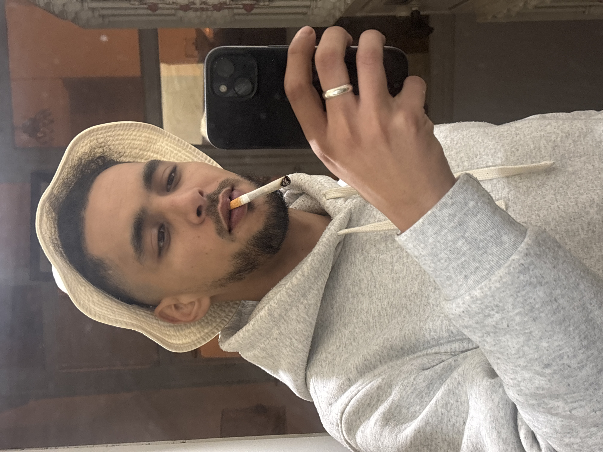 A male individual, appearing to be in his twenties, is depicted in a mirror selfie. He has dark hair with a fade on the sides, a short beard, and a mustache. He is wearing a light beige bucket hat and a light grey speckled hooded sweatshirt with cream drawstrings. A lit cigarette is held between his lips. He holds a dark-colored smartphone, featuring a triple-lens camera array, in his right hand, positioned to capture the reflection. A plain silver-colored band ring is visible on the ring finger of his right hand. The background, visible in the mirror's reflection, shows an interior space with warm, reddish-brown walls and a partial view of what appears to be a decorative light fixture or architectural element in the upper right. The scene is located in Tanger طنجة, Morocco.