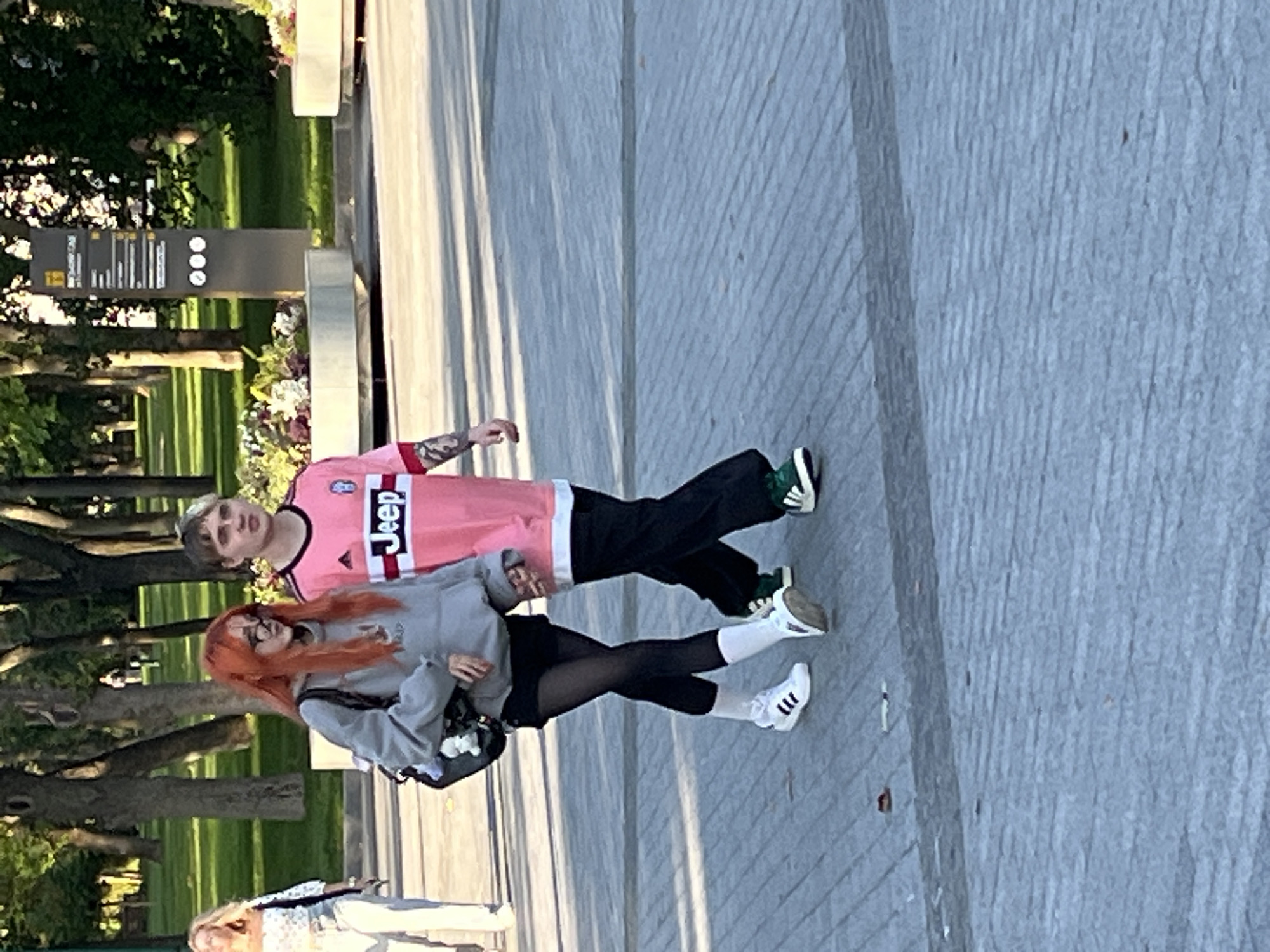 Two young adults, a woman with vibrant orange hair and glasses, and a man with blond hair, walk side-by-side on a paved walkway in a park. The woman wears a grey hoodie, black shorts, black tights, and white sneakers with black stripes. She carries a black backpack with a white plush toy attached. The man wears a bright pink t-shirt with "Jeep" written on it in white letters and black pants, along with dark green and black sneakers. They are walking from left to right in the frame, facing slightly towards the right. In the background, trees line the park, and a tall signpost with information is visible. The ground is paved with textured grey stones. Another woman in light-colored pants and a white patterned top is visible further to the left, walking away from the camera.