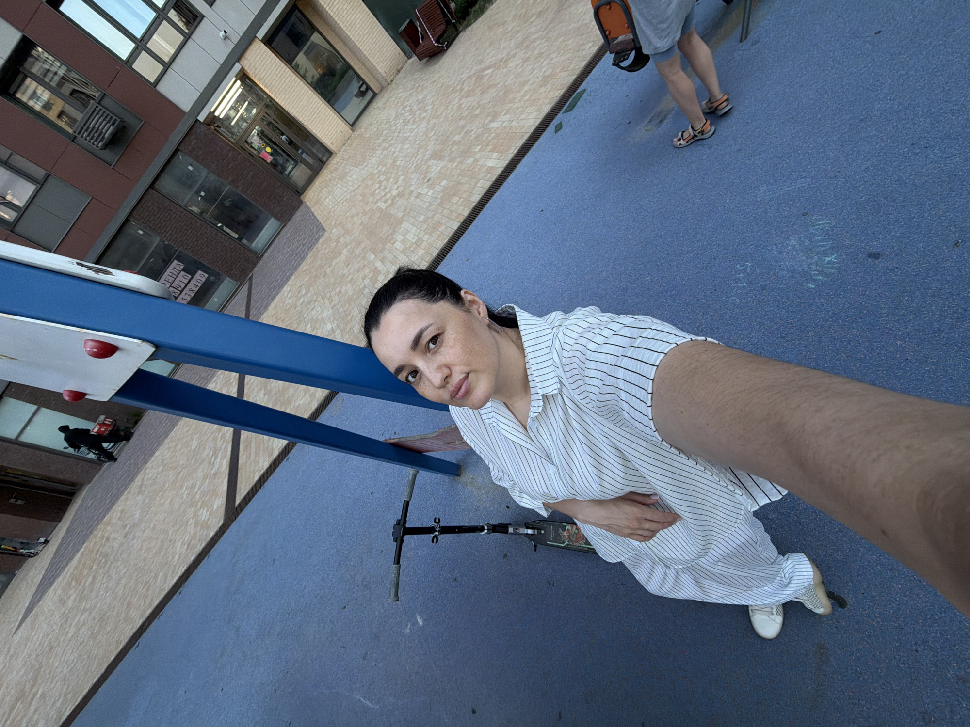 A high-angle, wide-angle selfie captures a woman standing on a blue playground surface next to a scooter. The woman, with dark hair pulled back and wearing a striped dress and white sneakers, is in the foreground, looking at the camera. Behind her, a person in a grey t-shirt and sandals is walking on a light-brown tiled surface. In the background, there is a modern building with numerous windows and a blue structure, possibly part of the playground. The scene appears to be in a courtyard or urban public space. The text "Аренда 8910538409" is visible on a window of the building.