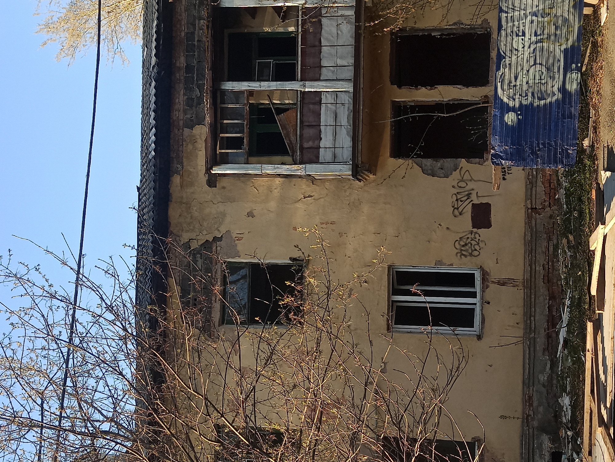 A weathered three-story building stands in an unknown location. The building's facade is a mix of crumbling stucco and exposed brick. Several windows are boarded up or broken, with a blue corrugated metal sheet covering one set. Graffiti is visible on the walls. A bare tree occupies the foreground, partially obscuring the building. The sky is visible in the upper left corner.
