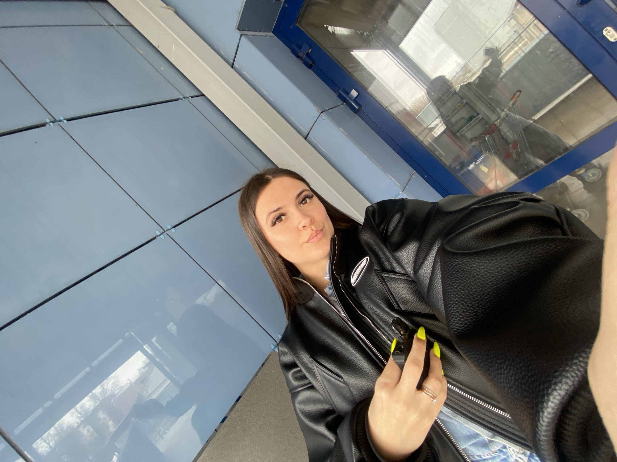 A woman is taking a selfie while standing outside a building in Chaikovsky, Russia. She is wearing a black leather jacket and has long dark hair. Her nails are painted bright yellow. In the background, there is a window reflecting a shopping cart and other items. The building exterior features blue panels and white framing.