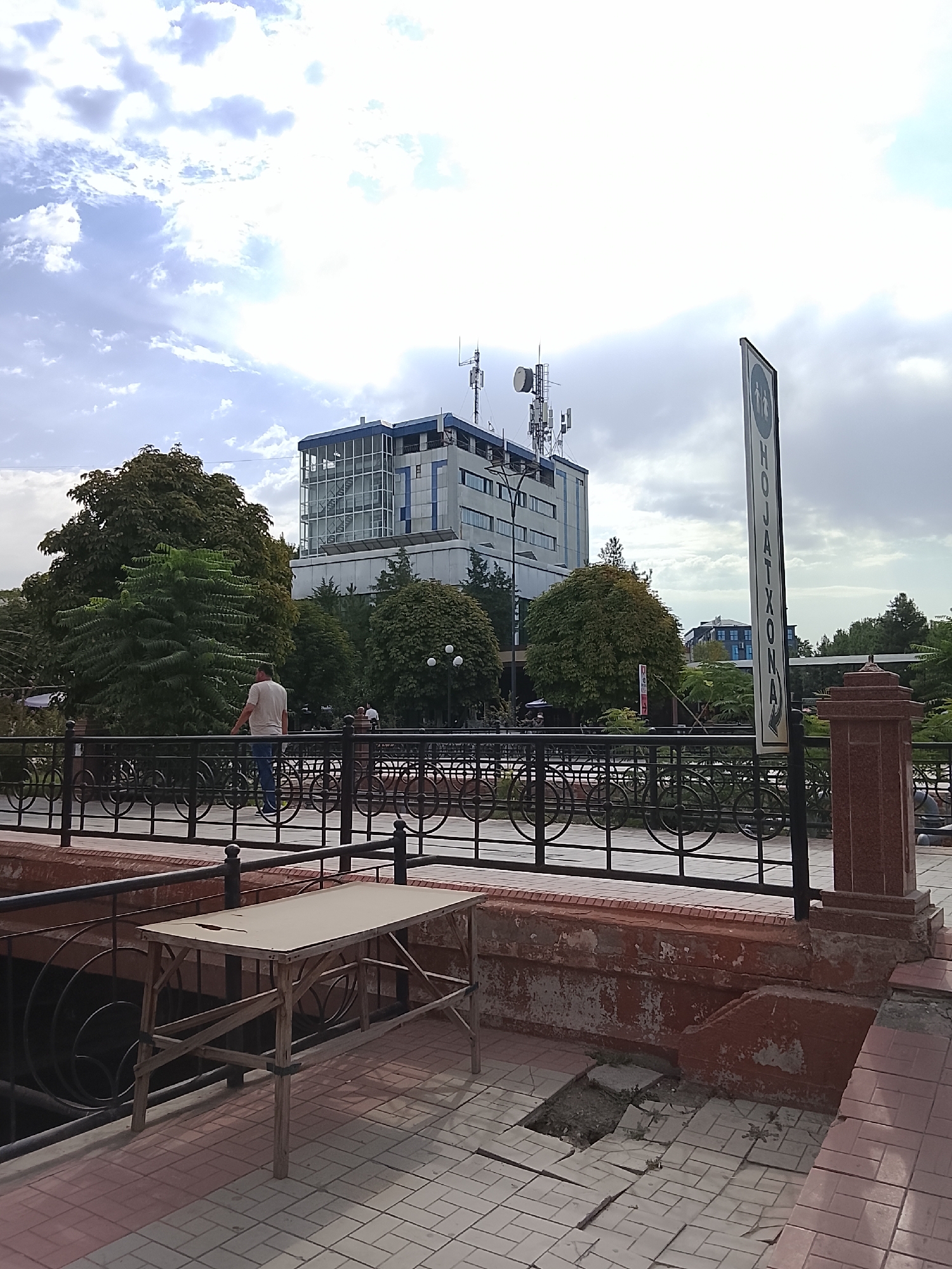 The image shows a view of a street in Fergana City, Uzbekistan, on a partly cloudy day. In the background, there is a modern, multi-story building with a blue-tinted glass facade and several communication antennas on its roof. Adjacent to the building is a cluster of large, mature trees with dense green foliage. In the foreground, a black metal fence with circular decorative elements runs horizontally across the scene. A light-colored wooden table with a worn surface stands near the fence. A person in a light-colored t-shirt and blue jeans is walking away from the camera on the other side of the fence. A vertical sign with Cyrillic lettering is visible on the right side of the image, partially obscured by the fence. The ground is paved with interlocking tiles. The overall impression is an urban environment with a mix of modern architecture and natural elements.