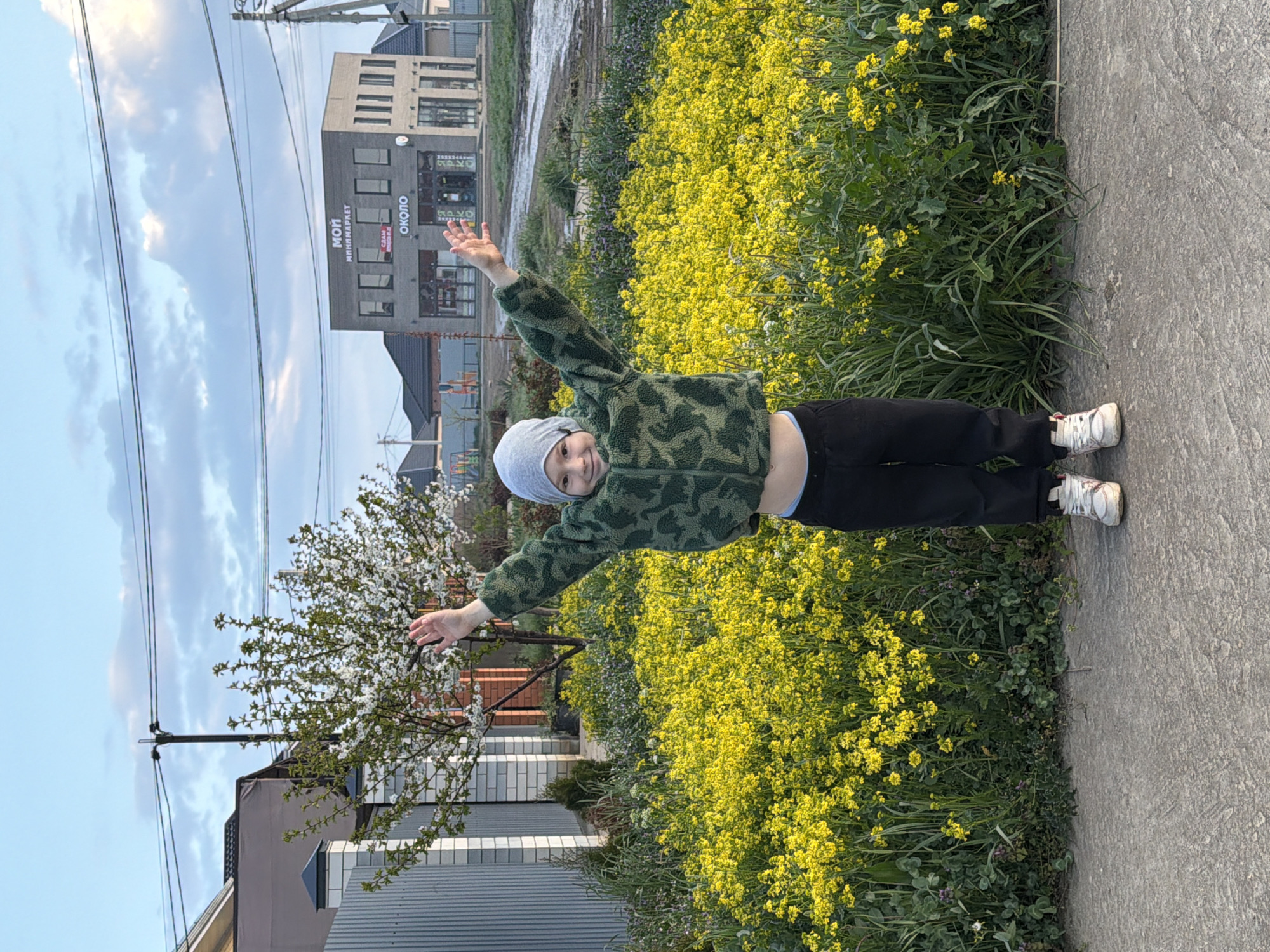 A child wearing a gray beanie, a green camouflage jacket, black pants, and white sneakers is standing on a concrete walkway in Rossiyskiy, Russia. The child has their arms outstretched. To the right of the child is a dense patch of yellow wildflowers. Behind the child, there is a white flowering tree, a fence, and a house. In the background, there is a multistory building with the Cyrillic inscription "МОЙ МИНИМАРКЕТ ОКОЛО." Overhead are electrical power lines extending into a blue sky.