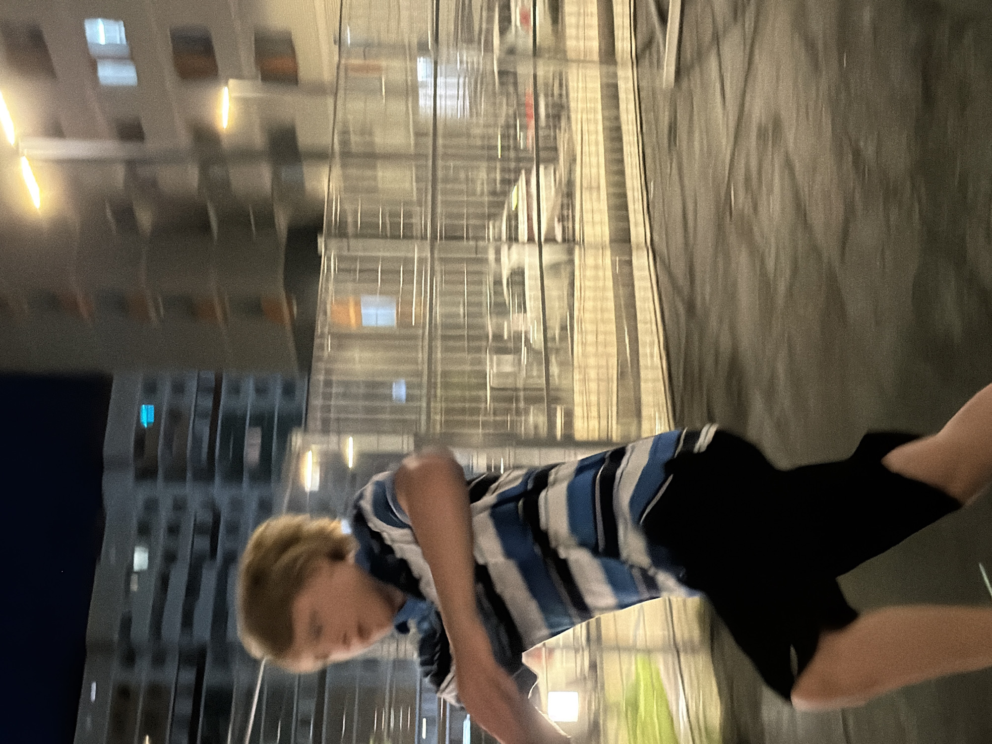 The image shows a young Caucasian boy with blond hair running outdoors at night. He is wearing a blue and white striped short-sleeved shirt and black shorts. The background consists of a blurred urban environment with tall apartment buildings and streetlights. There is a fence in the mid-ground with what appear to be cars visible through it. The ground is dark and wet, possibly asphalt. The lighting suggests artificial light sources. The overall impression is one of motion and a dimly lit, urban setting.