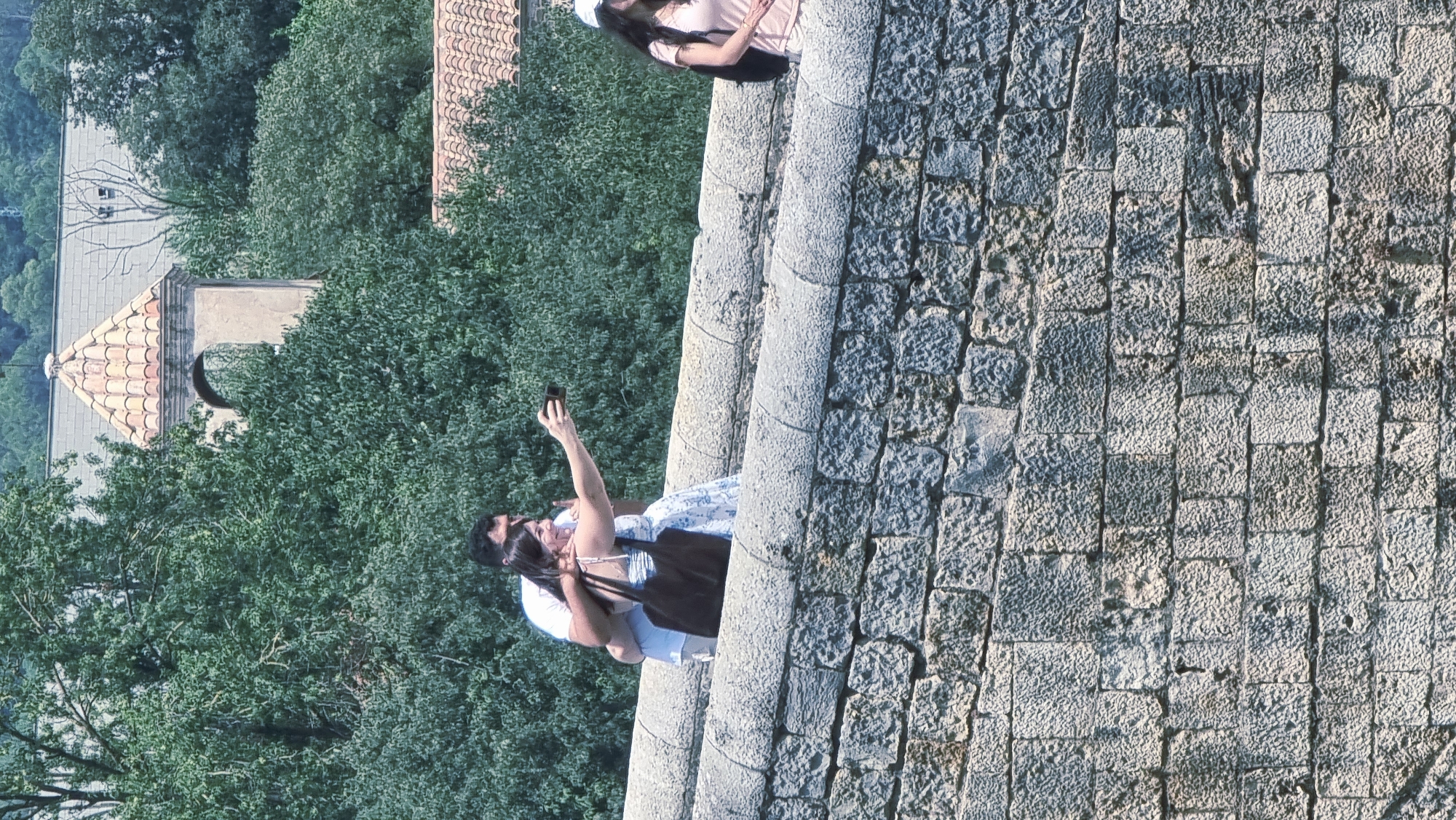 This image depicts a scene on a stone bridge in Besalú, Spain. In the foreground, a rough-hewn stone wall forms the base of the bridge. Above this wall, a wider, more finely constructed stone parapet lines the bridge's edge. Two people are visible on the bridge. A man, wearing a white t-shirt, has his arm around a woman. The woman, wearing a white and blue patterned dress, is holding a dark-colored tote bag over her shoulder and is in the process of taking a selfie with a mobile phone. Further along the bridge, to the right, a third person is seen from the side, wearing a pink t-shirt and a white cap, with a black backpack visible. The background is dominated by lush green trees, with a glimpse of a building featuring a tiled, conical roof and an arched opening visible through the foliage.