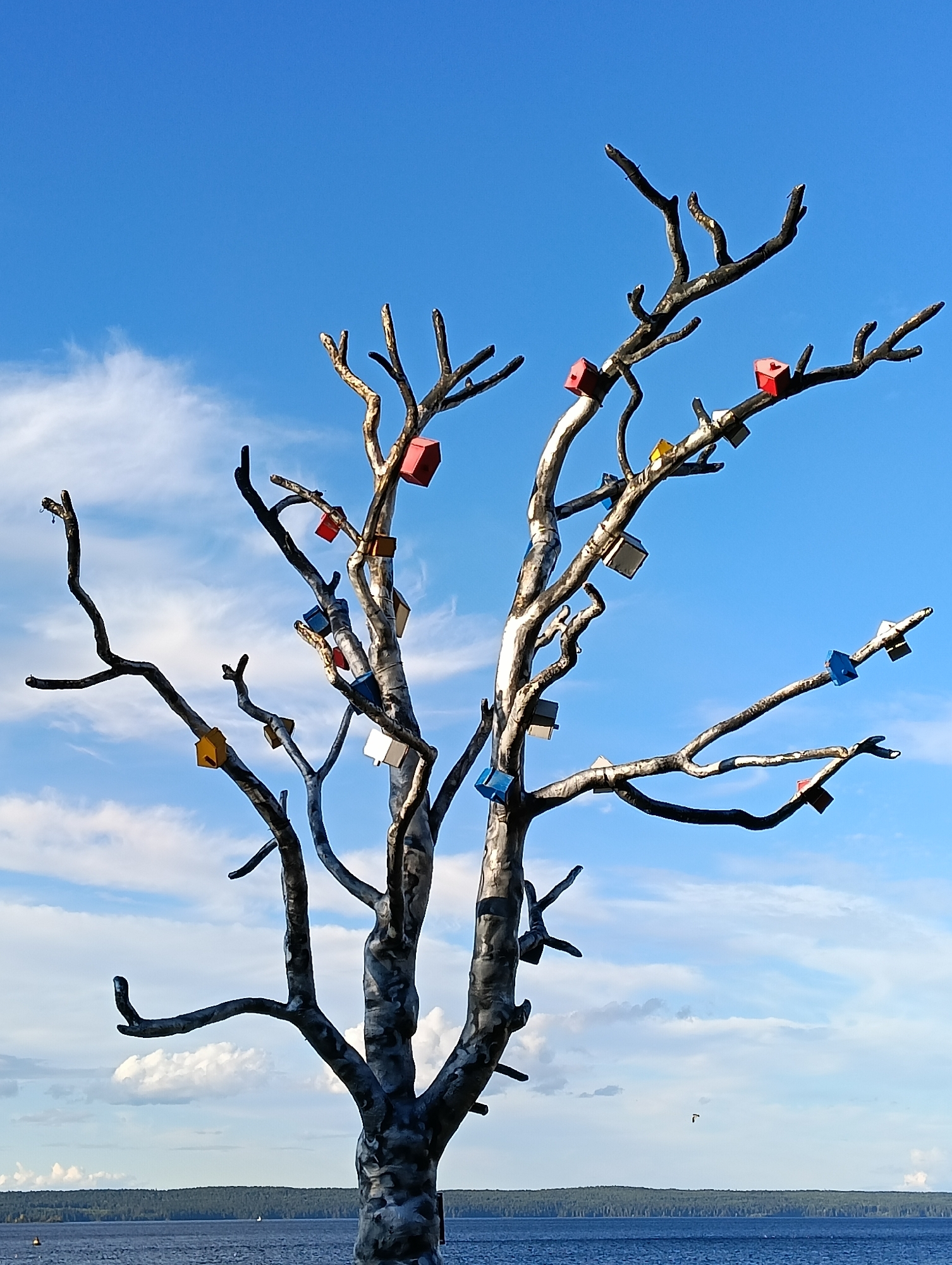 The image depicts a sculptural artwork of a bare tree with numerous small, colorful, cube-shaped structures attached to its branches. These structures resemble miniature houses or birdhouses and are painted in various bright colors, including red, blue, yellow, and white. The tree itself appears to be made of metal, with a textured, silvery-grey trunk and branches that are twisted and gnarled. The sculpture is situated outdoors, with a clear blue sky and scattered white clouds serving as the backdrop. In the lower part of the image, a body of water, likely a lake or bay, is visible, with a distant treeline on the horizon. This artwork is located in the city of Petrozavodsk, Russia, specifically on the Onega embankment.