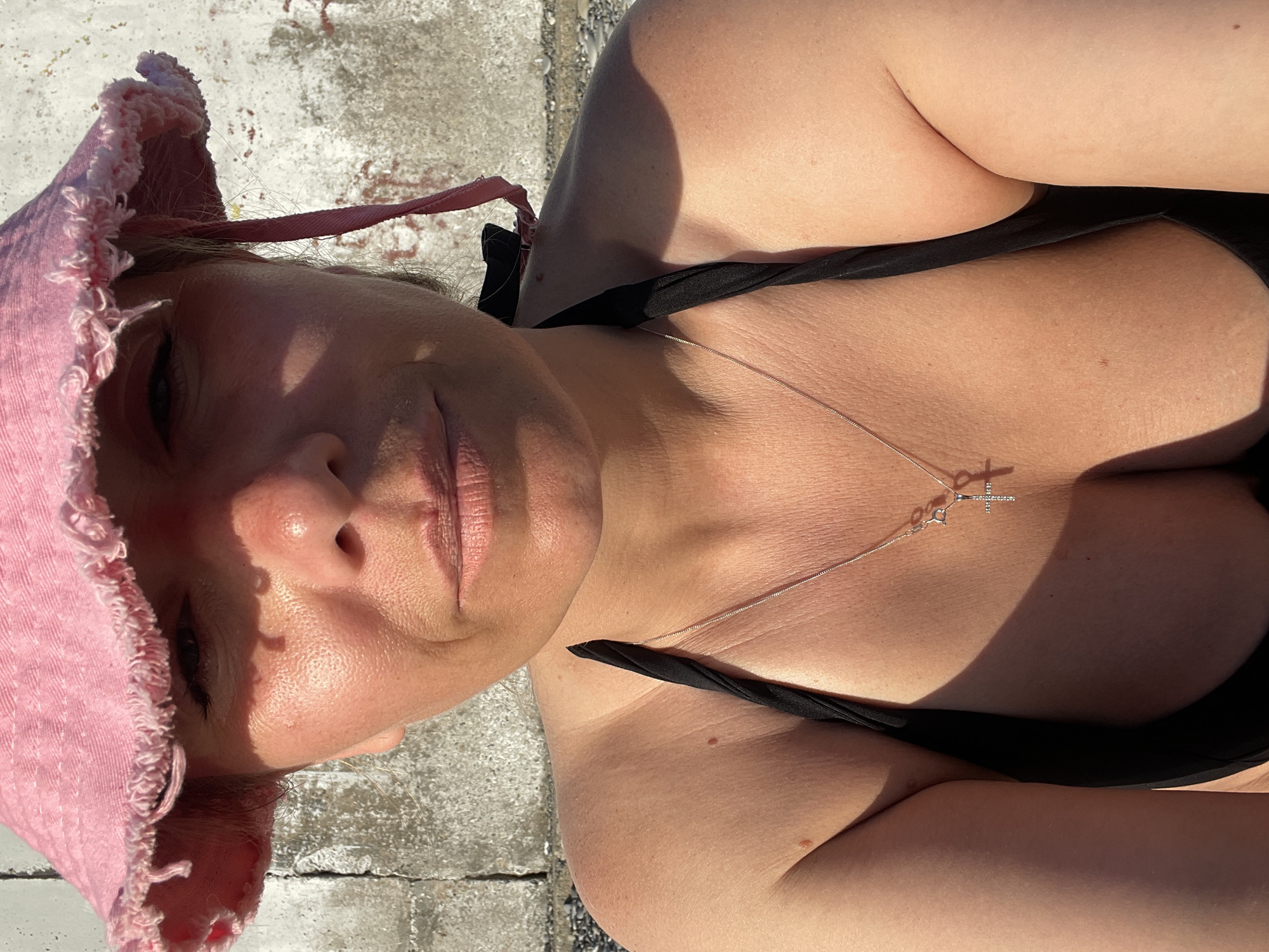 A close-up, chest-up, selfie-style photo shows a woman wearing a pink bucket hat and a black bikini top. She has a cross necklace on. The background is a textured concrete wall. The lighting suggests it is daytime, with shadows cast on her face and chest.