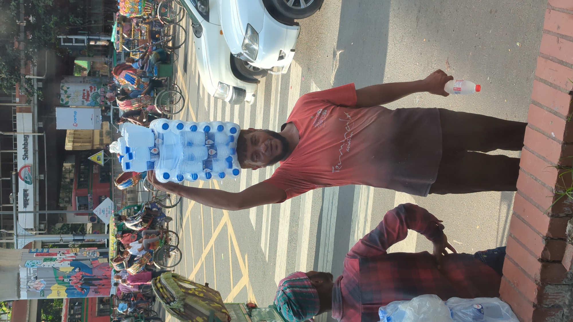 In Dhaka, Bangladesh, a man balances a stack of bottled water on his head, walking across a crosswalk. He wears an orange t-shirt and dark pants. Another man in a red plaid shirt and green and red head covering stands to his left. In the background, several rickshaws and cars are visible, along with an elevated railway station labeled "Shahbag." The environment is urban, with sunlight illuminating the scene.