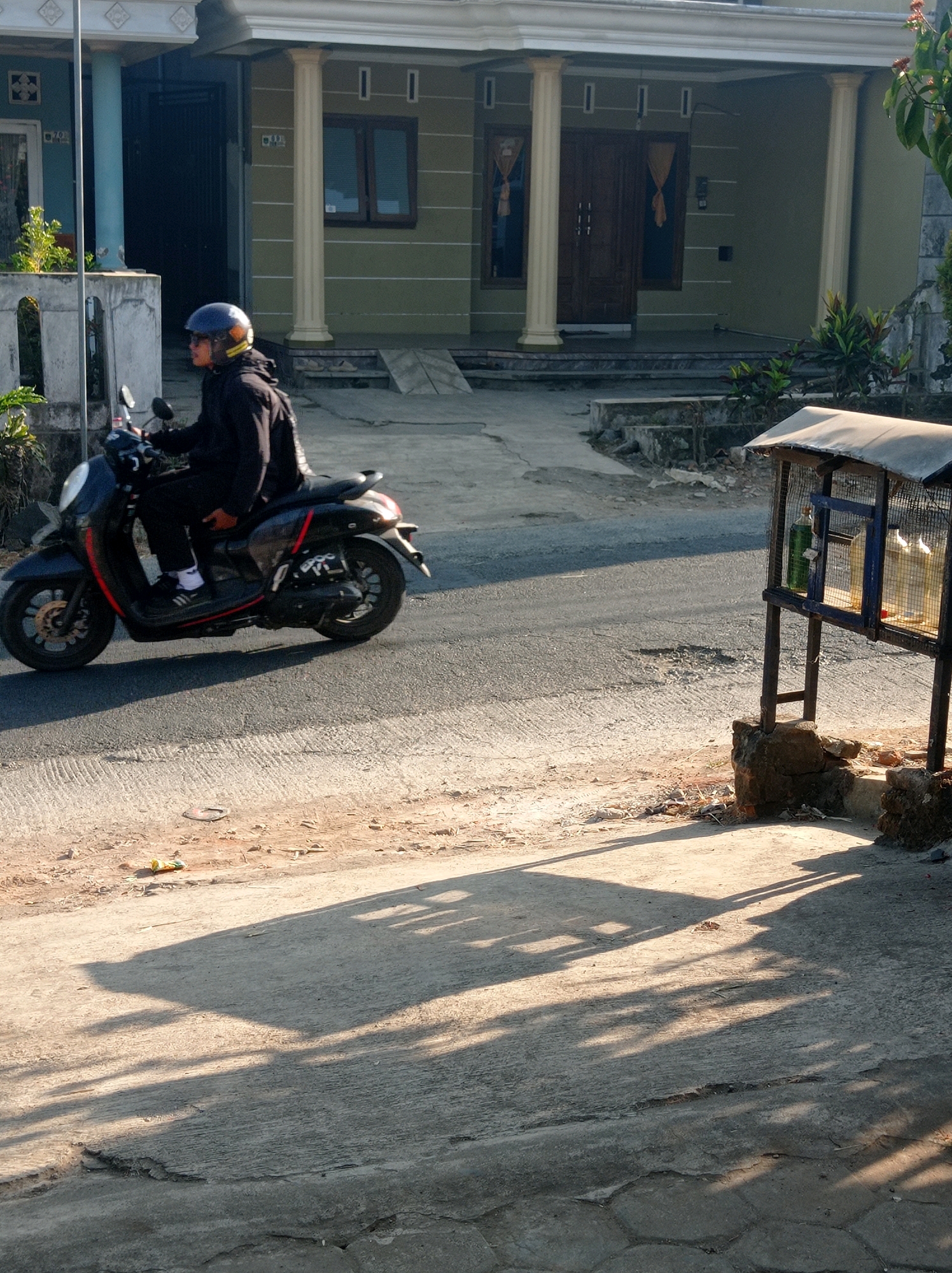 A person wearing a dark jacket and a blue helmet rides a dark-colored scooter with red accents. The scooter has a license plate that reads "EPOC YI". The person is riding on an asphalt road. In the background, there is a light green house with white pillars in front of the entrance. To the right of the road, there is a small, rustic wooden stand with a metal roof, containing several bottles of liquid. The environment appears to be a street in Purwosekar, Indonesia, during daylight, with long shadows cast on the ground.