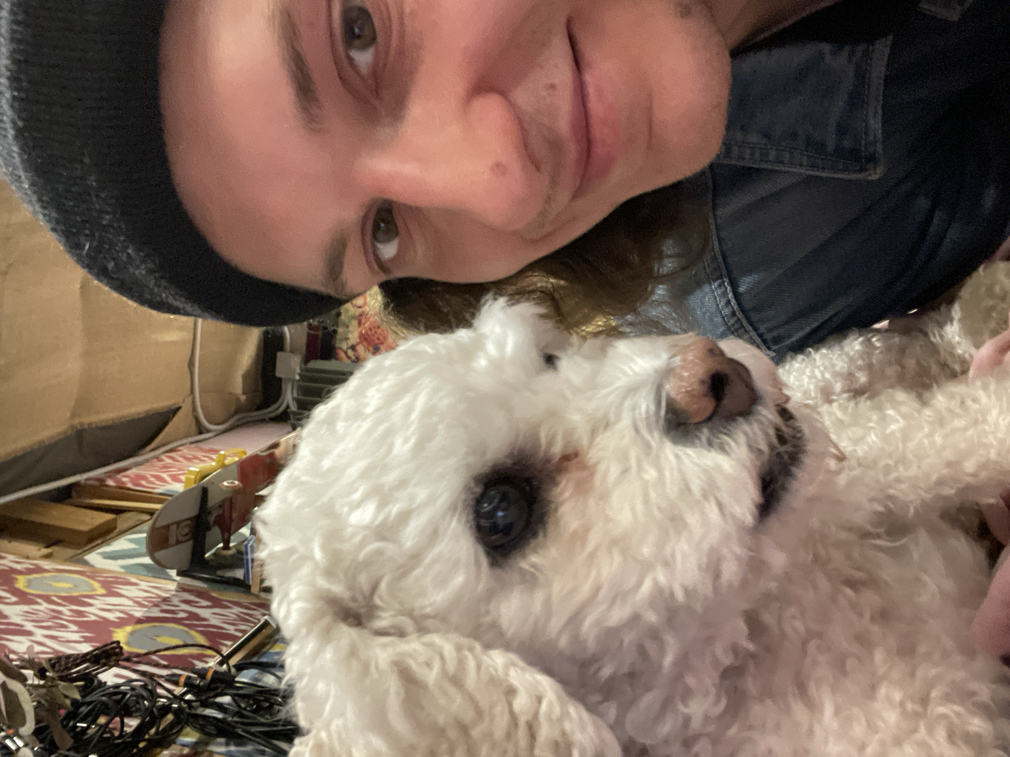 A close-up selfie shows a person wearing a dark beanie and a denim jacket, smiling slightly at the camera. A white, fluffy dog with dark eyes and a brown nose is positioned below the person's face, looking upwards. In the background, there is a colorful patterned rug, a cardboard box, electrical cords, and a toy airplane. The location is not discernible as being Yekaterinburg, Russia or any other specific city.
