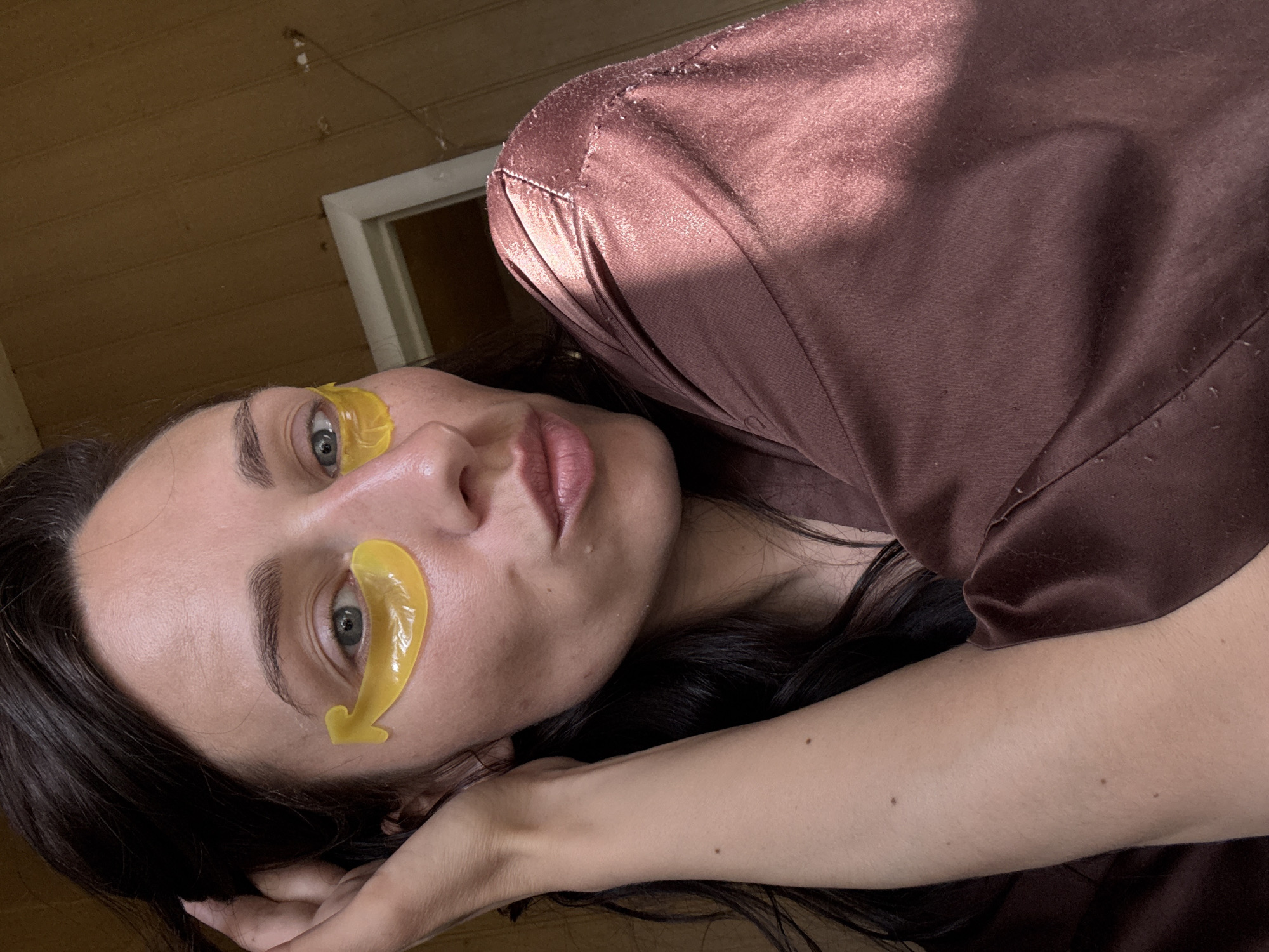 A woman with dark hair is reclining, wearing yellow eye masks. She has fair skin, gray eyes, and pink lips. She is wearing a dark brown satin-like garment. The background appears to be a room with wooden paneling and a doorway. There is no evidence to support the claim that the image was taken in Saint Petersburg, Russia.