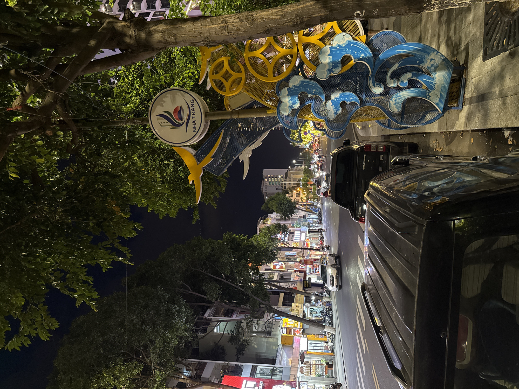 An eye-level, nighttime, medium shot captures a street scene in Nha Trang, Vietnam. The foreground is dominated by the roof of a black SUV, with a slightly dirty, textured surface. To the right of the SUV, a decorative metal sculpture featuring blue waves and yellow circular elements is positioned on the sidewalk, adjacent to a tree trunk. A street sign with the logo of Nha Trang and Vietnamese text is mounted on a pole, partially obscured by foliage.
The street is lined with buildings on the left, featuring multiple stories with balconies and illuminated windows. Various shops with signs in Vietnamese are visible along the storefronts. The road is paved and has white lane markings. Several vehicles, including cars and motorbikes, are present on the street, some in motion and others parked. Streetlights and building lights illuminate the scene, creating a vibrant nighttime atmosphere. Trees with dark green foliage line the sidewalk, their branches extending over the street. The sky is dark, indicating it is nighttime. The overall environment suggests a busy urban street with commercial activity.