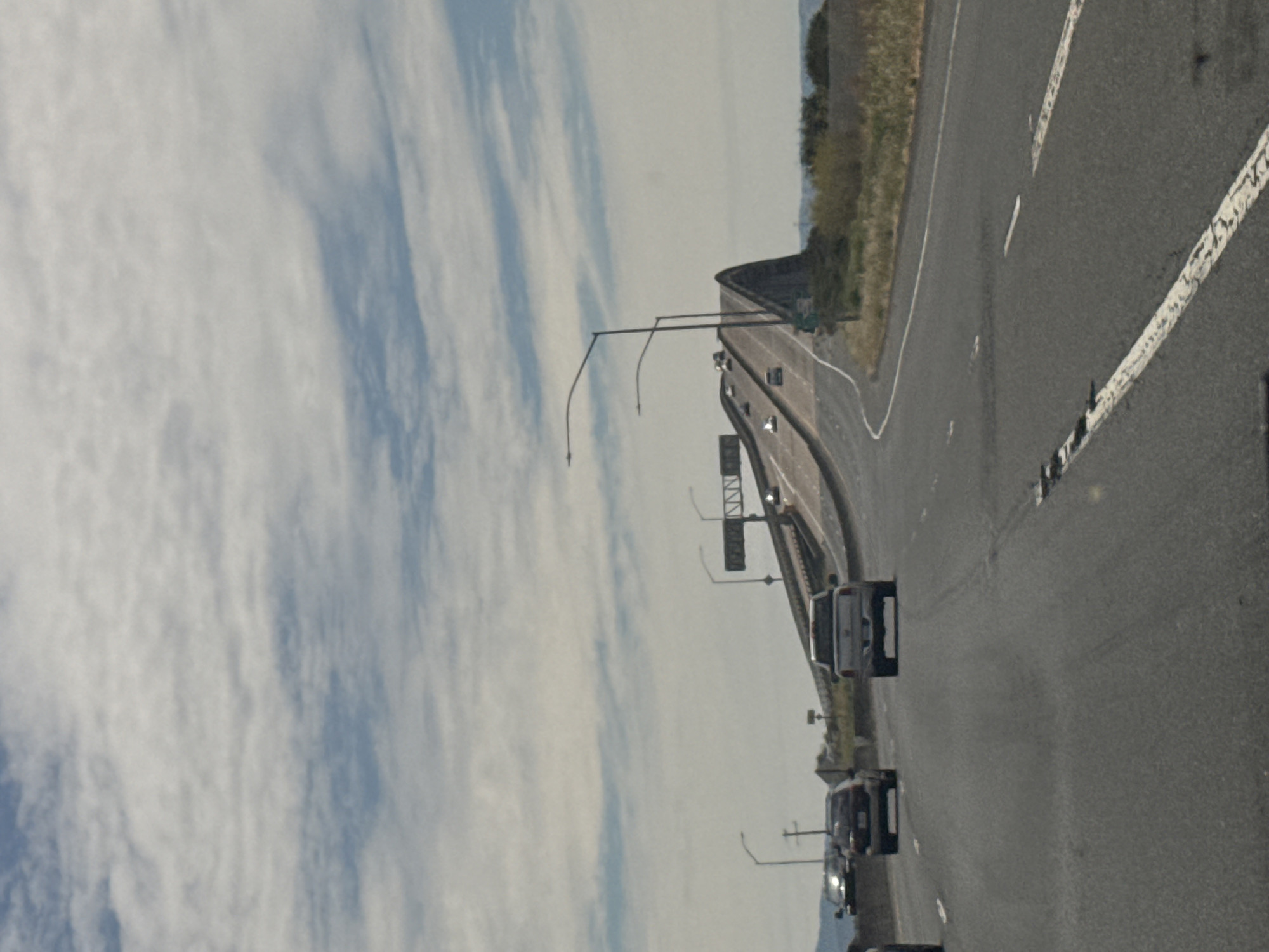 The image depicts a wide, multi-lane highway leading to a high bridge with a distinctive arch. The sky is partly cloudy, with white clouds spread across a blue expanse. The bridge appears to be concrete and steel, with traffic signs visible at its peak. Cars are traveling on the highway, including a silver pickup truck in the foreground and several other vehicles further ahead. The surrounding landscape consists of dry, grassy hills and sparse vegetation. The scene is captured from the perspective of someone in a vehicle on the highway, looking forward towards the bridge. The overall impression is of a vast, open highway infrastructure under a bright, partly cloudy sky. The location is identifiable as the Carquinez Bridge in Vallejo, California, United States of America, given its unique arch design and the surrounding topography.