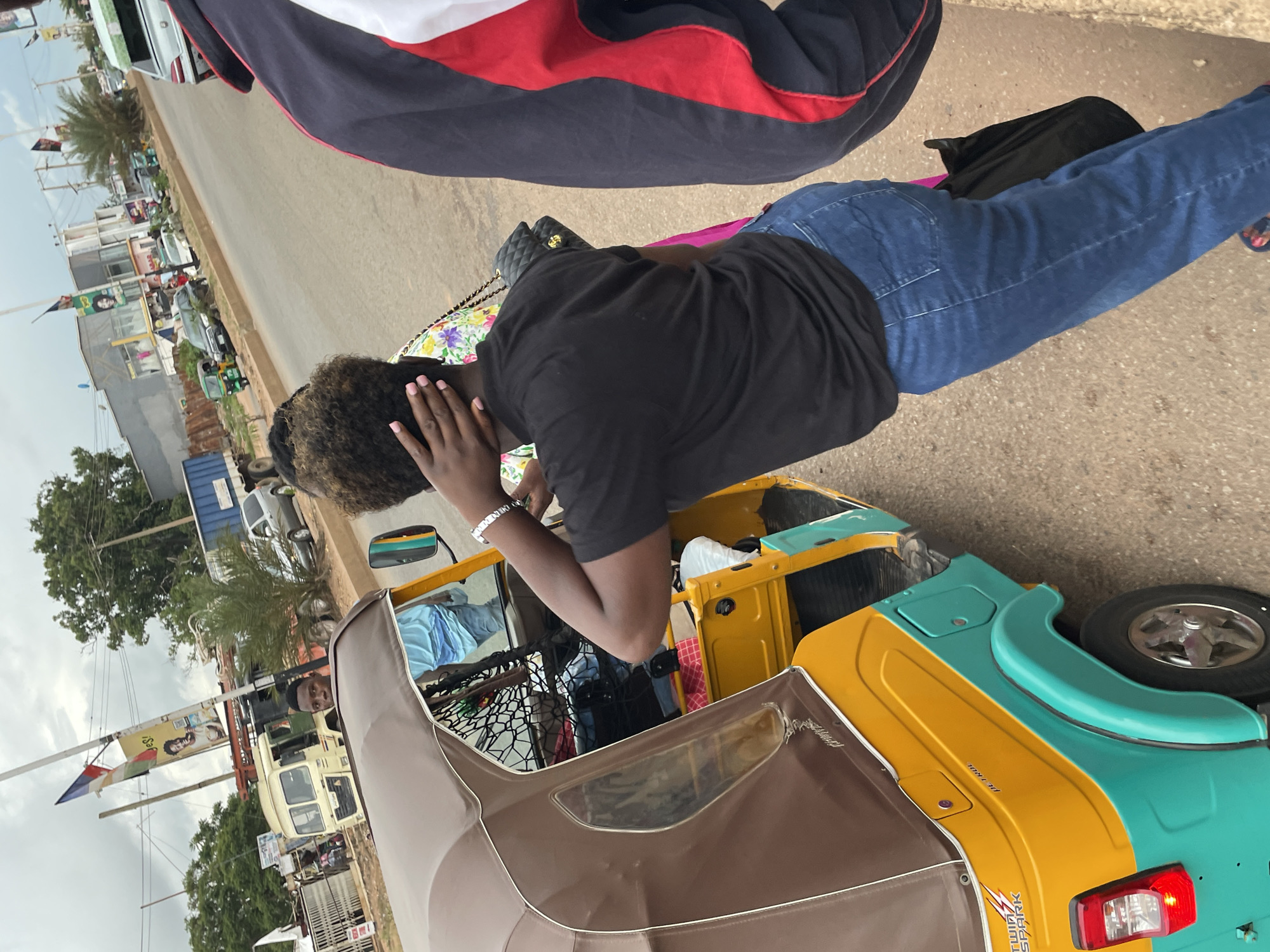 A medium shot shows a woman with curly dark hair wearing a black t-shirt and blue jeans. She is standing near the back of a yellow and turquoise auto-rickshaw, touching her neck with her left hand. To her right, a person in a dark jacket with red and white stripes is partially visible. The auto-rickshaw is parked on a paved road in an urban environment. In the background, there are buildings, trees, a utility pole with flags and signage, and other vehicles including a truck and a silver car. The sky is overcast. The scene appears to be in Jos, Nigeria, based on the presence of auto-rickshaws, the architectural style of the buildings, and the general streetscape. The overall impression is of a typical street scene in a Nigerian city.