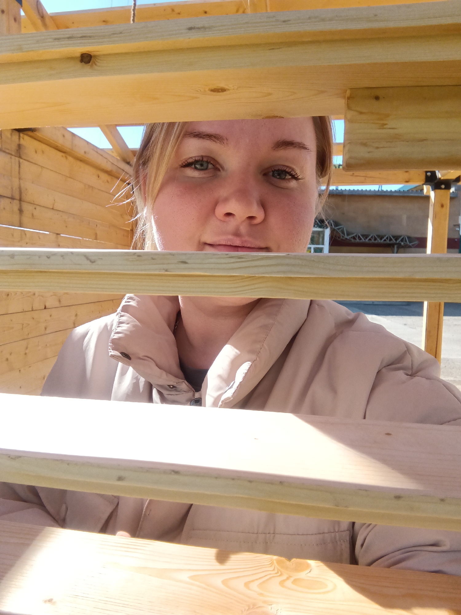 A woman is seen through the wooden slats of a structure, likely under construction, in Фоки, Russia. The woman has blonde hair pulled back, fair skin, and is wearing a beige jacket. The structure appears to be made of light-colored wood, possibly pine. In the background, a building and outdoor features are visible, suggesting an urban or suburban environment. The scene is captured in daylight.