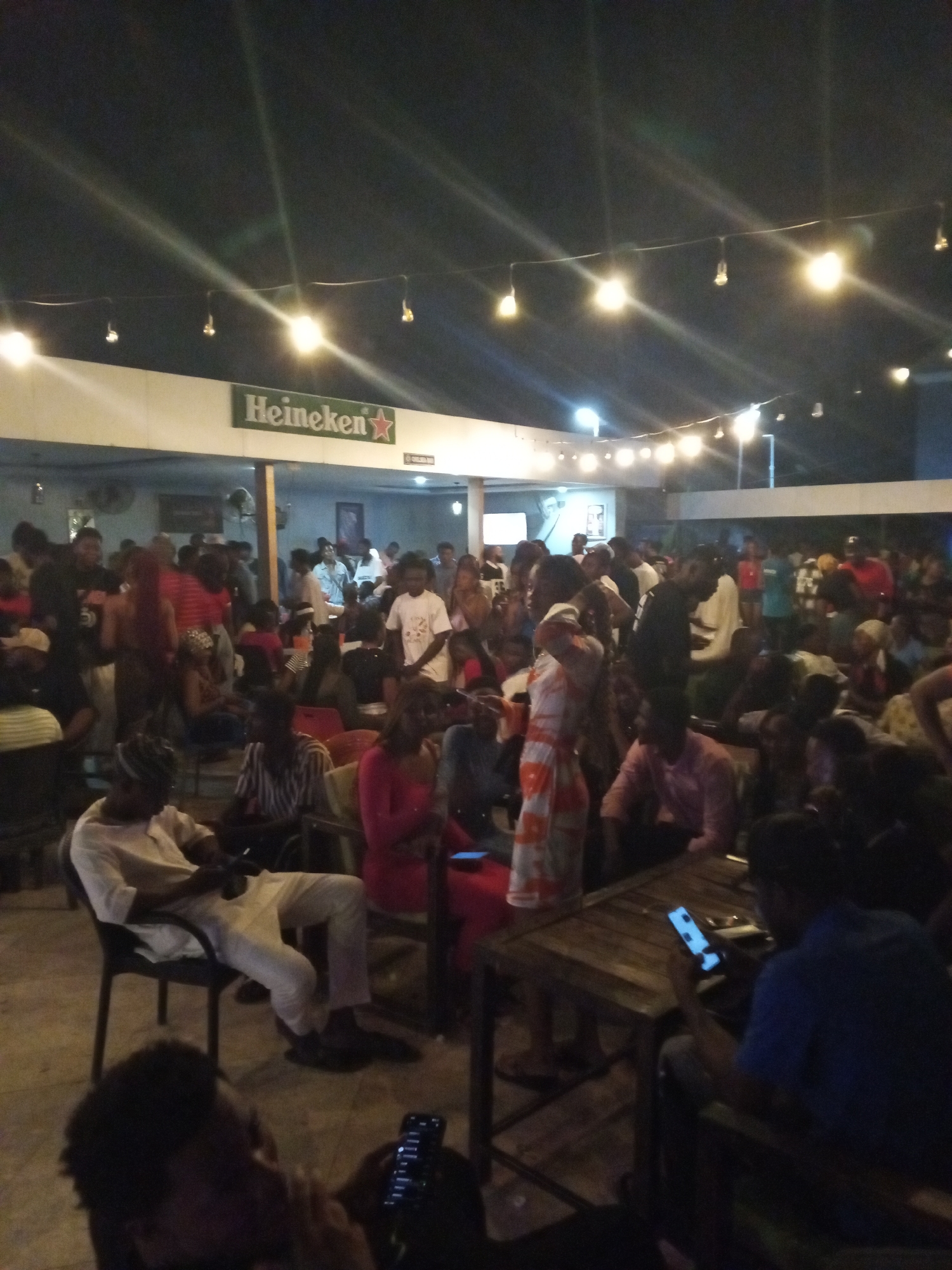The image depicts a crowded outdoor gathering at night in Adankolo, Nigeria. The location is likely a bar or event space with a "Heineken" sign visible. Numerous people are present, both standing and seated at tables. Many individuals are using mobile phones. The area is lit by strung-up light bulbs, providing ambient illumination.