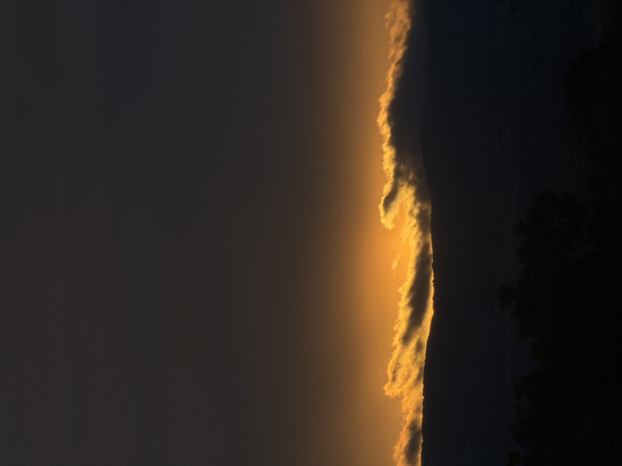 A nighttime photograph captures a dark, silhouetted landscape under a sky illuminated by a sliver of orange light near the horizon. The scene is dominated by the deep blacks of a mountain range and scattered trees in the foreground. Above the mountains, a band of clouds is brightly lit from below by a setting or rising sun, creating a dramatic contrast against the darkening sky. The image appears to be taken from an elevated viewpoint, looking across the landscape. No specific identifiable landmarks of Blagoevgrad, Bulgaria, are visible.
