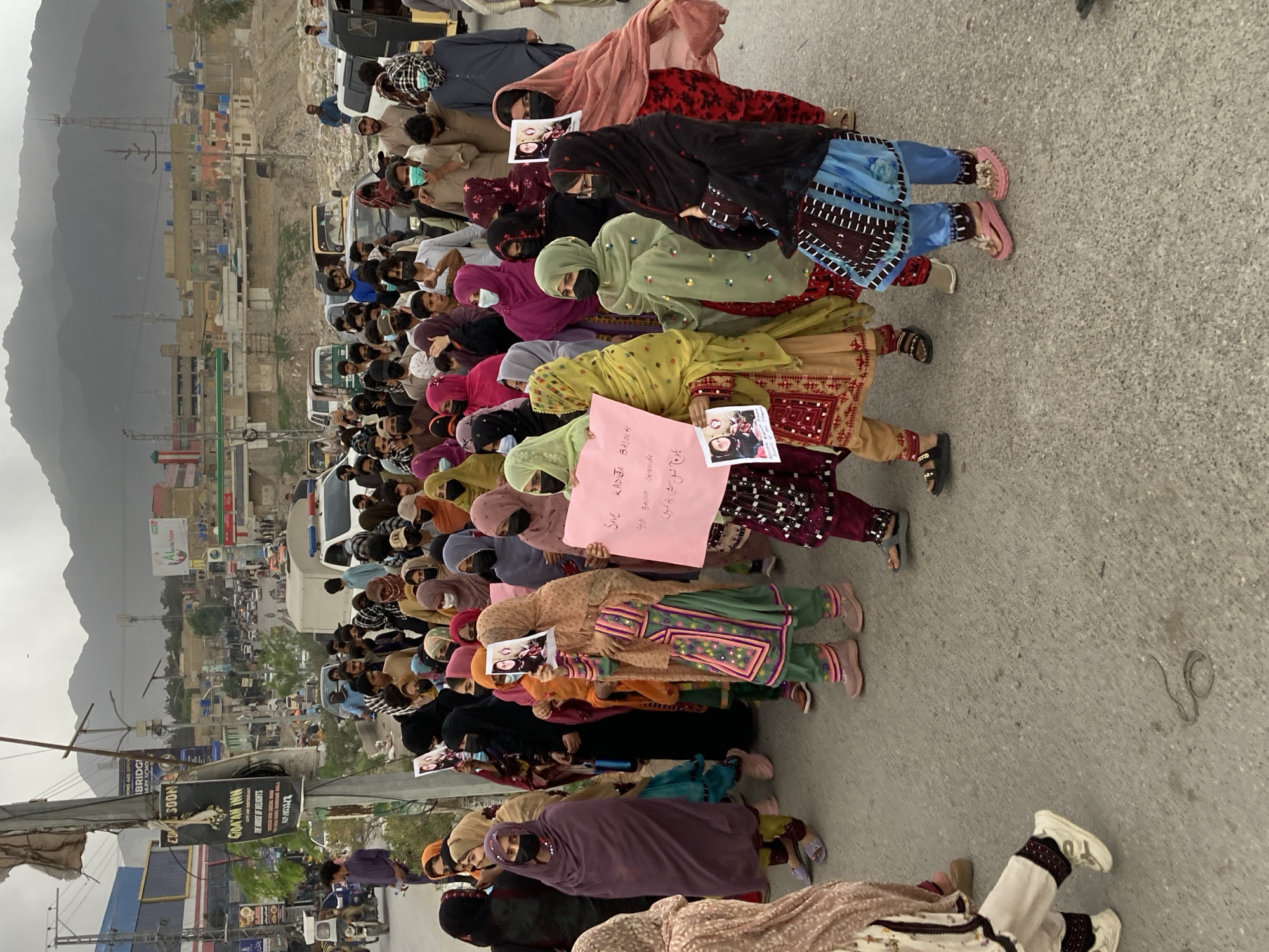 A ground-level, medium-shot photograph shows a line of women demonstrating in Quetta, Pakistan. The group of approximately fifty women kneel or sit in a line on a paved road, facing uphill toward the photographer. The women wear traditional Baloch clothing, including long, colorful dresses and headscarves. Most of the women wear black face masks. Some women hold signs, including pink signs with text in English and Urdu that read "Save Kadiga Baloch, Stop Baloch Genocide." Many of the women are holding laminated photos of two young women.
Behind the women, a crowd of men watches the demonstration. Vehicles, including cars and trucks, are partially visible between the women and the men. The town is visible in the background. Buildings appear to be tightly packed together. Mountains loom in the distance behind the town. Utility poles and wires are visible along the road. Signs for local businesses are visible, including "Gida'an Inn" and "Bons are open."