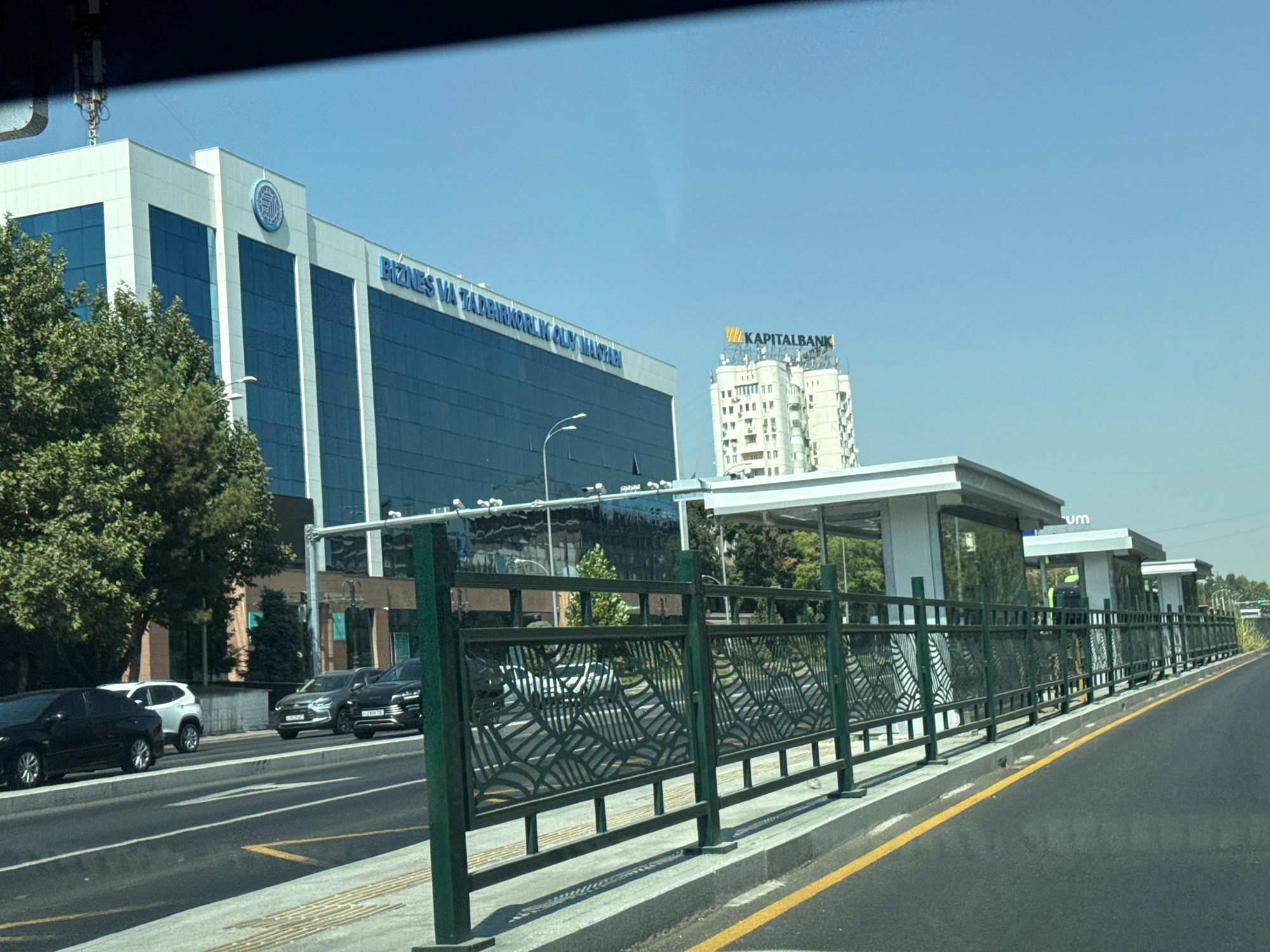 In Tashkent, Uzbekistan, a wide, asphalt road runs parallel to a modern bus stop with a decorative green metal fence and an adjacent sidewalk. Behind the bus stop, a large, multi-story building with blue-tinted glass windows and white paneling is visible. The building has "BIZNES VA TADBIRKORLIK OLIM MANTABI" inscribed in blue lettering. Further in the background, another taller white building with a sign that reads "KAPITALBANK" stands against a clear blue sky. Several cars, including a black sedan, a silver SUV, and a white SUV, are present on the road. The scene is captured from the perspective of someone inside a vehicle, looking out through a window. The overall environment suggests an urban setting on a clear day.