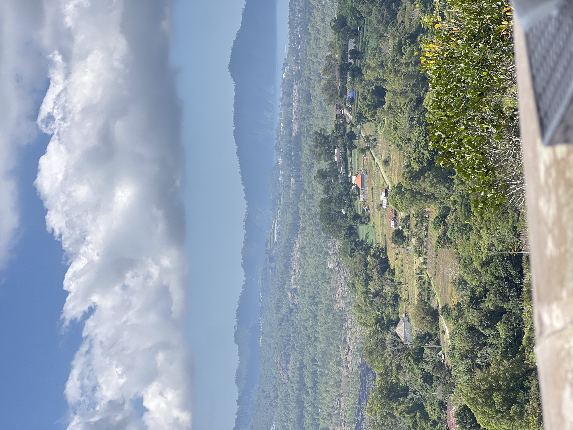 The image captures a sweeping panoramic view of the Kintamani region in Bali, Indonesia. The foreground is dominated by a blurred, stone-like structure, possibly a balcony or ledge, with a portion of a woven outdoor seating surface visible on the right. Beyond this, the landscape unfolds in layers of lush green vegetation, with dense trees and some scattered patches of dry grass and cultivated fields. Several small houses with red and orange roofs are nestled within the greenery, connected by a winding dirt road.
Further into the mid-ground, the terrain becomes more rugged, with rocky outcrops and a denser forest canopy. In the distance, a body of water, likely Lake Batur, is visible, shimmering under the bright sky. Beyond the lake, rolling hills and mountains form the horizon, shrouded in a soft, bluish haze. The sky above is a vibrant blue, punctuated by large, dramatic white clouds that cast soft shadows on the landscape below. The overall impression is one of a serene, rural mountainous landscape bathed in the bright light of day.
