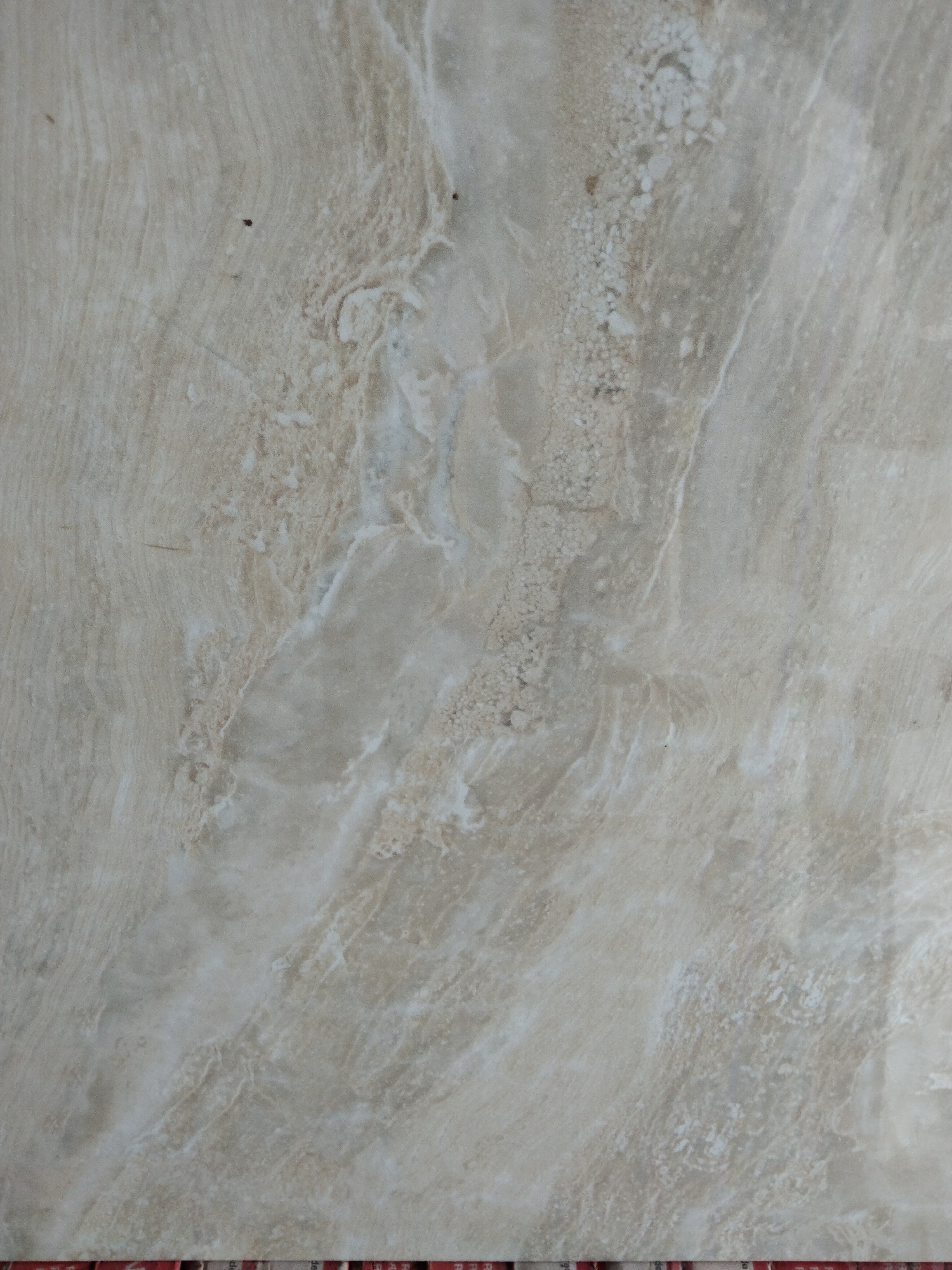 The image displays a close-up, overhead view of a beige-colored tile with a marbled texture. The tile features variations in tone, with lighter beige swirls and darker, grayish-brown veins running through it, resembling natural stone. There are a few small, dark specks on the surface, possibly dirt or imperfections. The lighting appears to be even, highlighting the subtle textures and patterns of the tile. The bottom edge of the image shows a portion of what appears to be printed paper or cardboard with some red and white markings, suggesting the tile might be part of a display or packaging. There is no visible indication of its location in Ciseeng, Indonesia, as the image exclusively focuses on the tile itself.