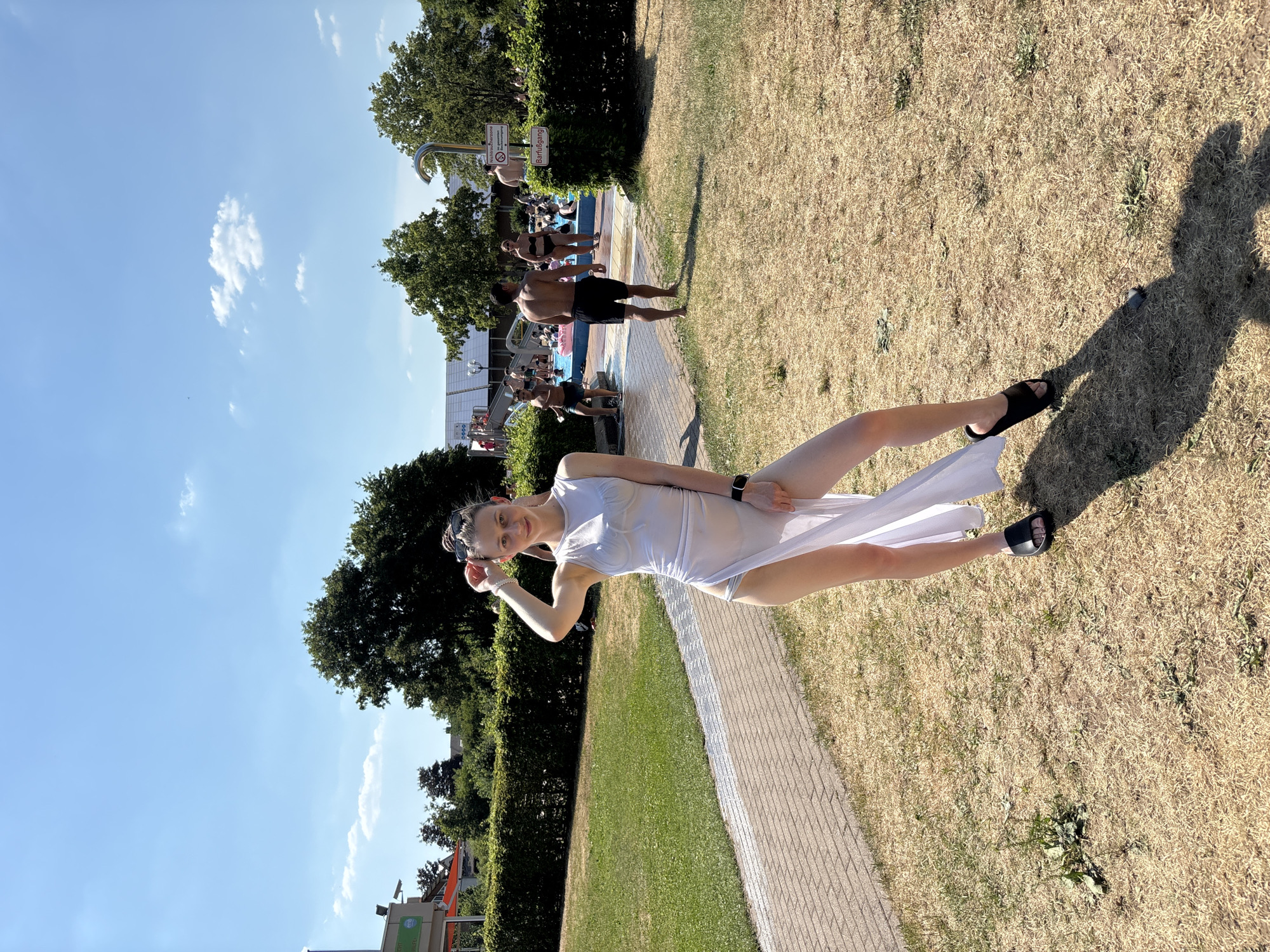 In Bürgstadt, Germany, a young woman in a flowing white dress poses by a paved path, her legs extended and a playful smile on her face. She is barefoot on one foot and wearing a black sandal on the other, with a smartwatch on her wrist. The dress, sheer and light, is cinched at the waist and drapes elegantly to the ground. The setting appears to be a public outdoor space, possibly a park or a recreational area, with a grassy lawn on one side and a path on the other.
Behind the woman, a swimming pool area is visible, with people in swimwear lounging and walking around. The water is a vibrant blue, reflecting the clear, sunny sky. Lush green trees line the background, casting shadows on the dry, sun-baked grass. The overall atmosphere is one of a warm, sunny day, likely in the summer, with people enjoying leisure time outdoors. The clear blue sky dotted with a few wispy clouds suggests fair weather. There is a sign visible near the pool area with German text, and another sign with text that includes "Barfußgang", meaning "barefoot path". The overall mood is relaxed and cheerful, capturing a moment of summer enjoyment.