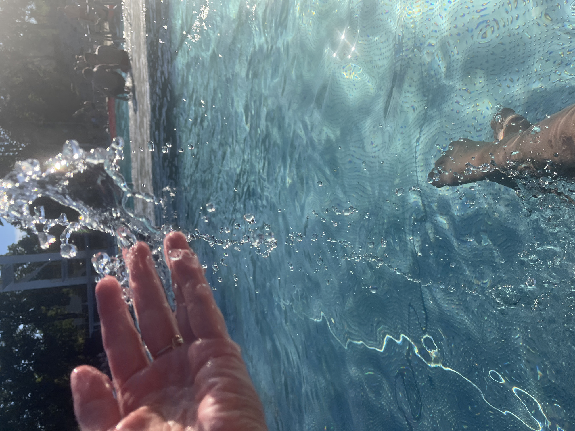 A first-person perspective shot captures a hand splashing water in a pool. The hand, with a ring on one finger, is in the upper left of the frame, with water droplets cascading from it. The pool water is a vibrant blue, with light reflecting off the surface and creating shimmering patterns on the pool floor. In the background, other people are visible, some sitting on the pool deck and others in the water. The overall setting appears to be a public swimming pool on a sunny day.