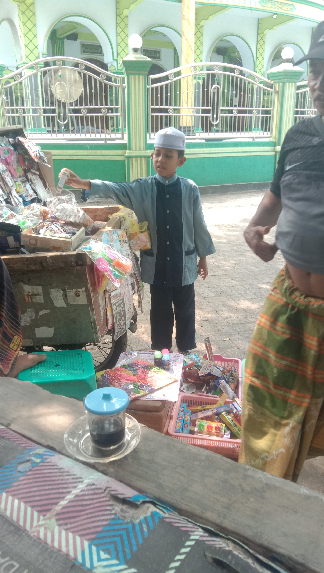A young boy, wearing a white peci and a grey and black patterned tunic with dark pants, stands near a cart filled with various items. He holds a small, white-packaged item in his right hand, seemingly offering it or showing it to someone. The cart is laden with colorful merchandise, including toys and candies, and appears to be a mobile stall. In the foreground, a glass of dark liquid sits on a saucer on a wooden surface. To the right, an adult, partially visible, wears a striped sarong. The background features a building with green and white architectural details and arches, suggestive of a mosque, possibly located in Ngempit, Indonesia. The ground is paved, and the overall scene depicts a street-side commerce setting.