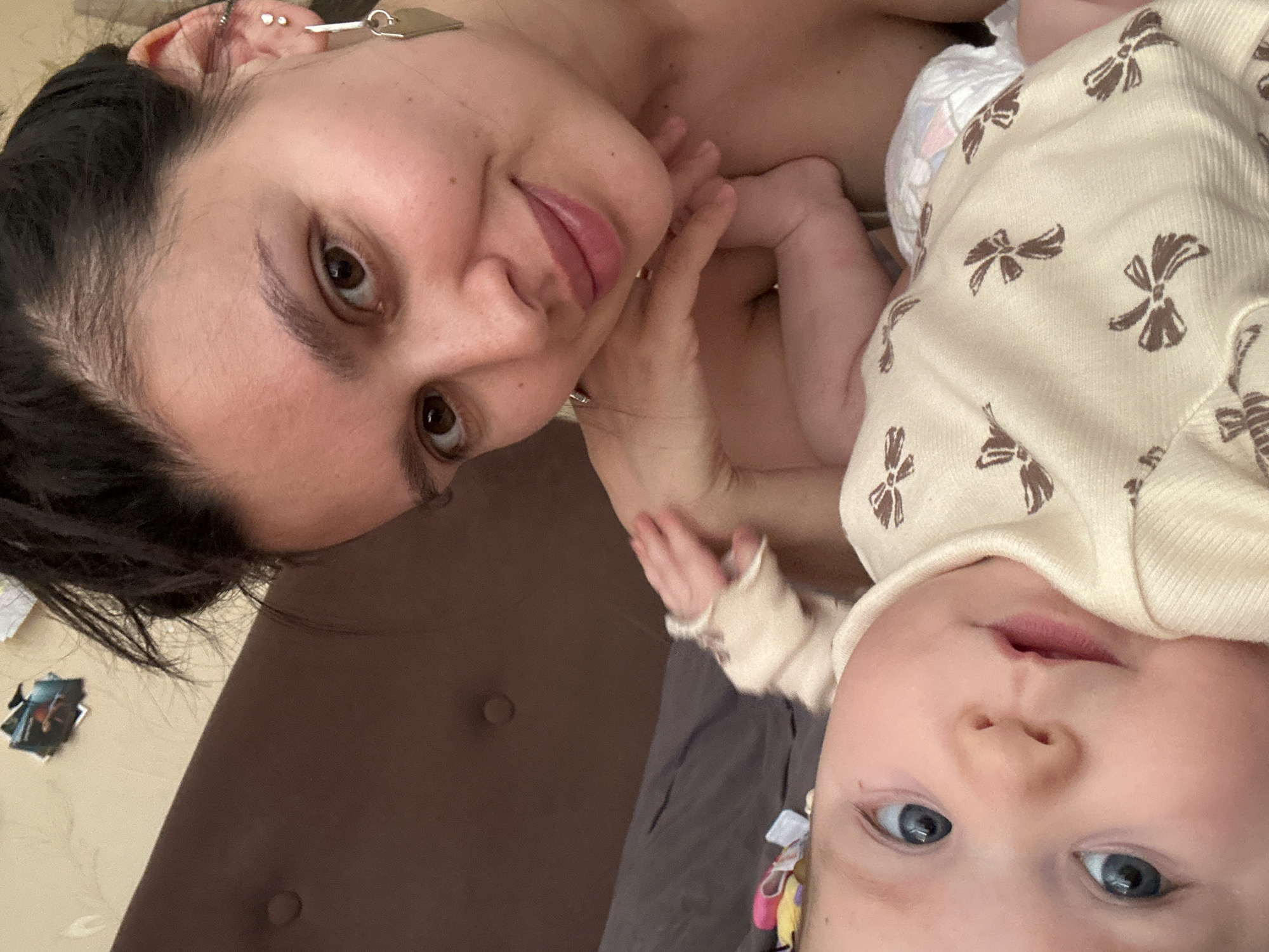 A woman and baby are posing for a close-up selfie, rotated counter-clockwise. The woman has dark hair, dark eyes, and is wearing multiple earrings in her left ear. She is touching her face with her right hand. The baby is wearing a light-colored onesie with brown bowties and has blue eyes. The background includes a brown headboard and gray bedding. It is unclear if the picture was taken in the city of Chaikovsky, Russia.