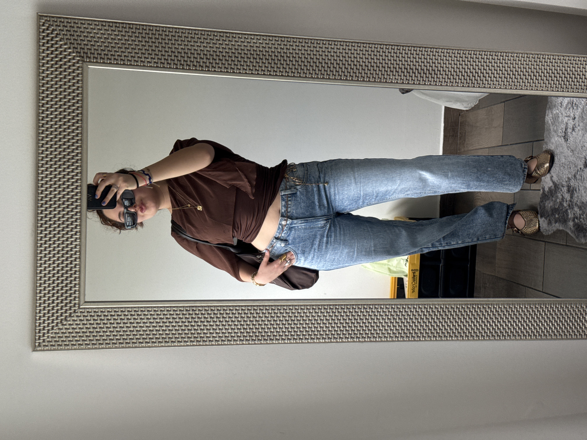 A full-body selfie is taken in front of a large, horizontally oriented mirror with a patterned frame, presumably indoors. The subject, a woman with brown hair and sunglasses, holds a smartphone in her right hand to capture the image. She wears a brown long-sleeve top, blue jeans with a chain detail at the waist, and gold-colored flats. Her left hand rests on her hip. The background reflected in the mirror shows a plain white wall, a tiled floor, a grey rug, and some items including a container with a yellow label ("solo.Orient") visible in the reflection. Based on the items visible in the reflection, the location appears to be a domestic interior, likely a residence in Mexico City, Mexico.