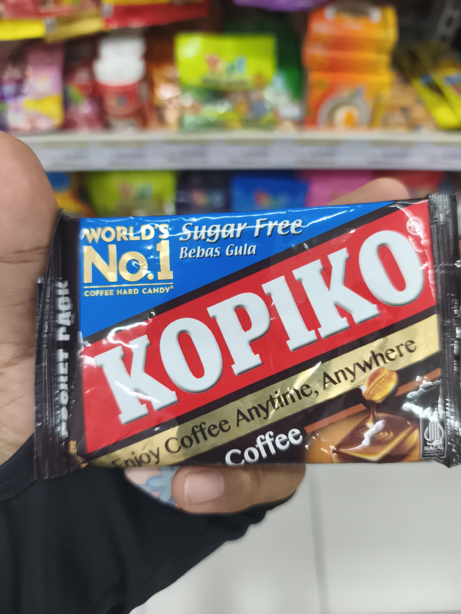 An individual's hand is holding a package of Kopiko coffee hard candy in the foreground. The candy wrapper is primarily blue, red, and gold, featuring the brand name "KOPIKO" in large white letters. Text on the wrapper includes "WORLD'S Sugar Free No.1 Bebas Gula" and "COFFEE HARD CANDY®". A tagline below the brand name reads "Enjoy Coffee Anytime, Anywhere". A small Halal certification logo is visible on the right side of the wrapper. The background shows a blurred retail environment, likely a convenience store or supermarket aisle, with shelves stocked with various colorful products. The scene appears to be located in Kemiri Kidul, Indonesia, as indicated by the Halal certification logo which specifies "INDONESIA". The lighting suggests indoor illumination typical of a commercial establishment.