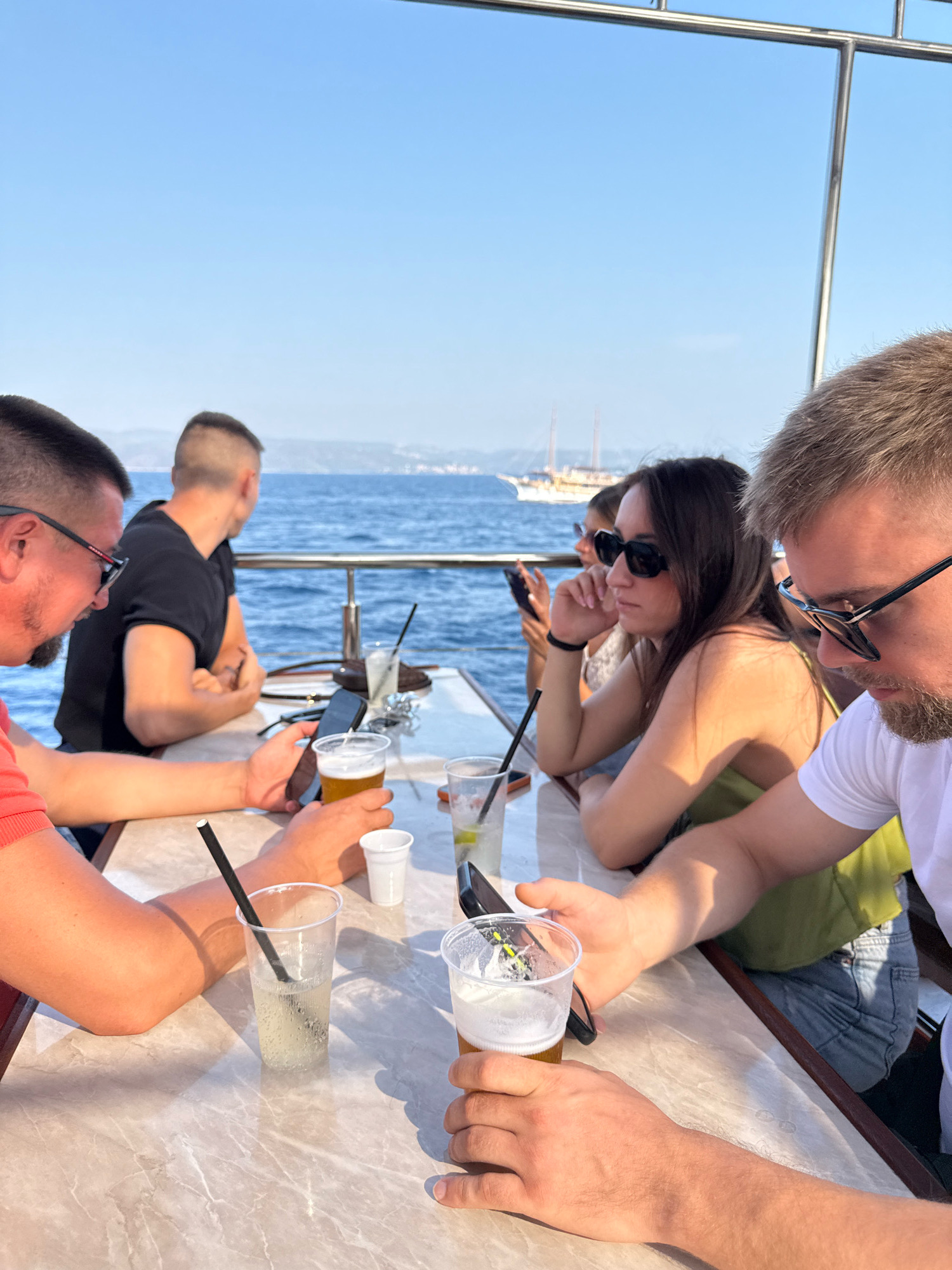 The image shows a group of people sitting at a table on a boat in the sea near the city of Selca, Croatia. The sky is clear and blue, and the water is calm. The table is made of marble and has several drinks and cell phones on it. There are at least four people visible in the image, all appearing to be adults. Two men are in the foreground, one on the left wearing a coral-colored shirt and sunglasses, and another on the right wearing a white t-shirt and sunglasses. A woman with long brown hair is seated between them, wearing a green top and sunglasses. Another person, a man with short dark hair and a black t-shirt, is seated on the left side of the table, facing away from the camera. The man on the right is holding a cell phone and a glass of beer. The man on the left is also holding a cell phone. In the background, there are some sailboats and the coastline of the city of Selca is visible. The overall atmosphere appears to be relaxed and social.