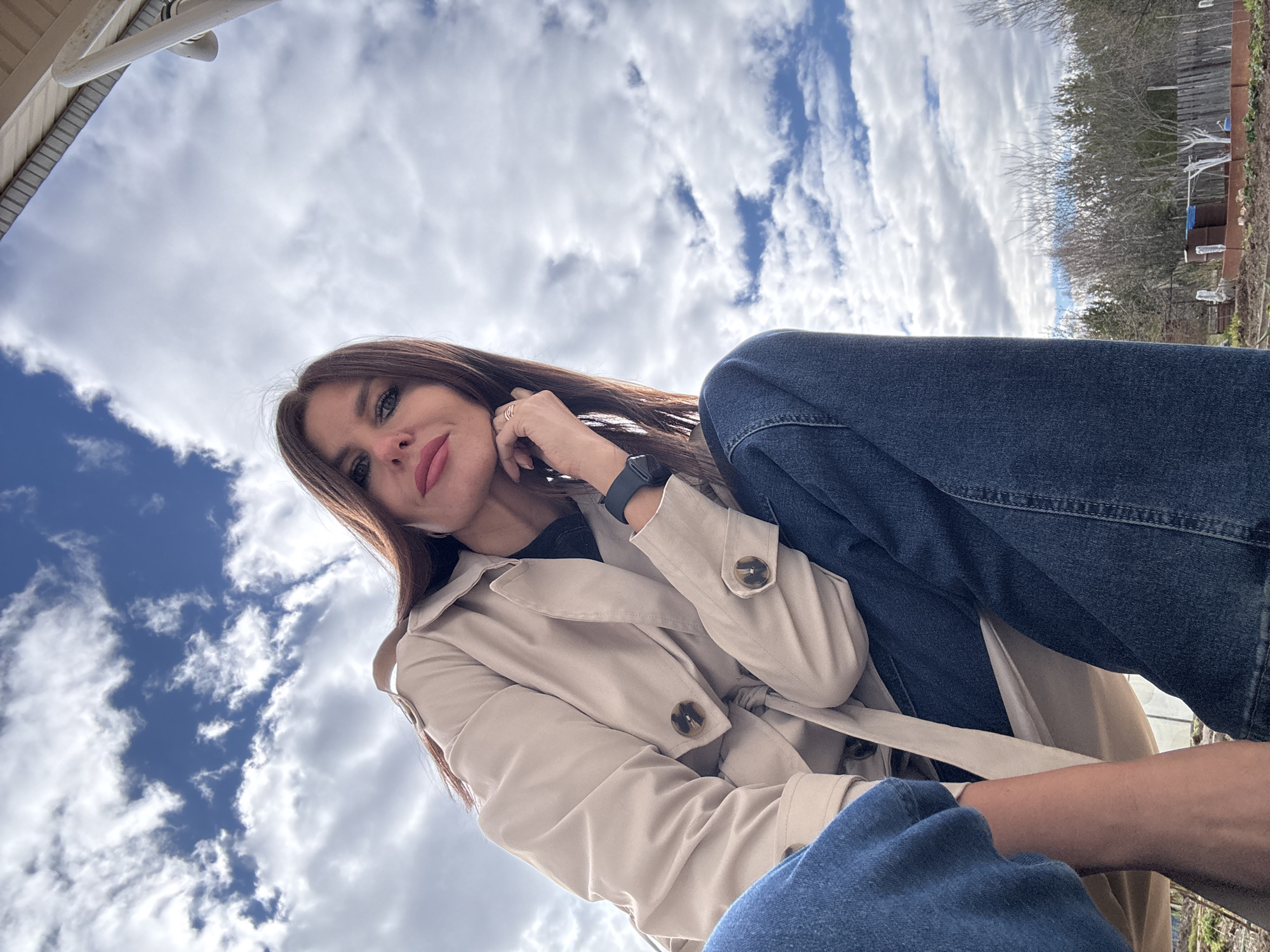 A woman is depicted in an upward-angled selfie. She has long brown hair and is wearing a light tan trench coat and blue jeans. She has a wristwatch on her left wrist. The background shows a sky filled with white and blue clouds. In the upper right of the image, some trees and structures are visible. The location is Dubovaya, Russia.
