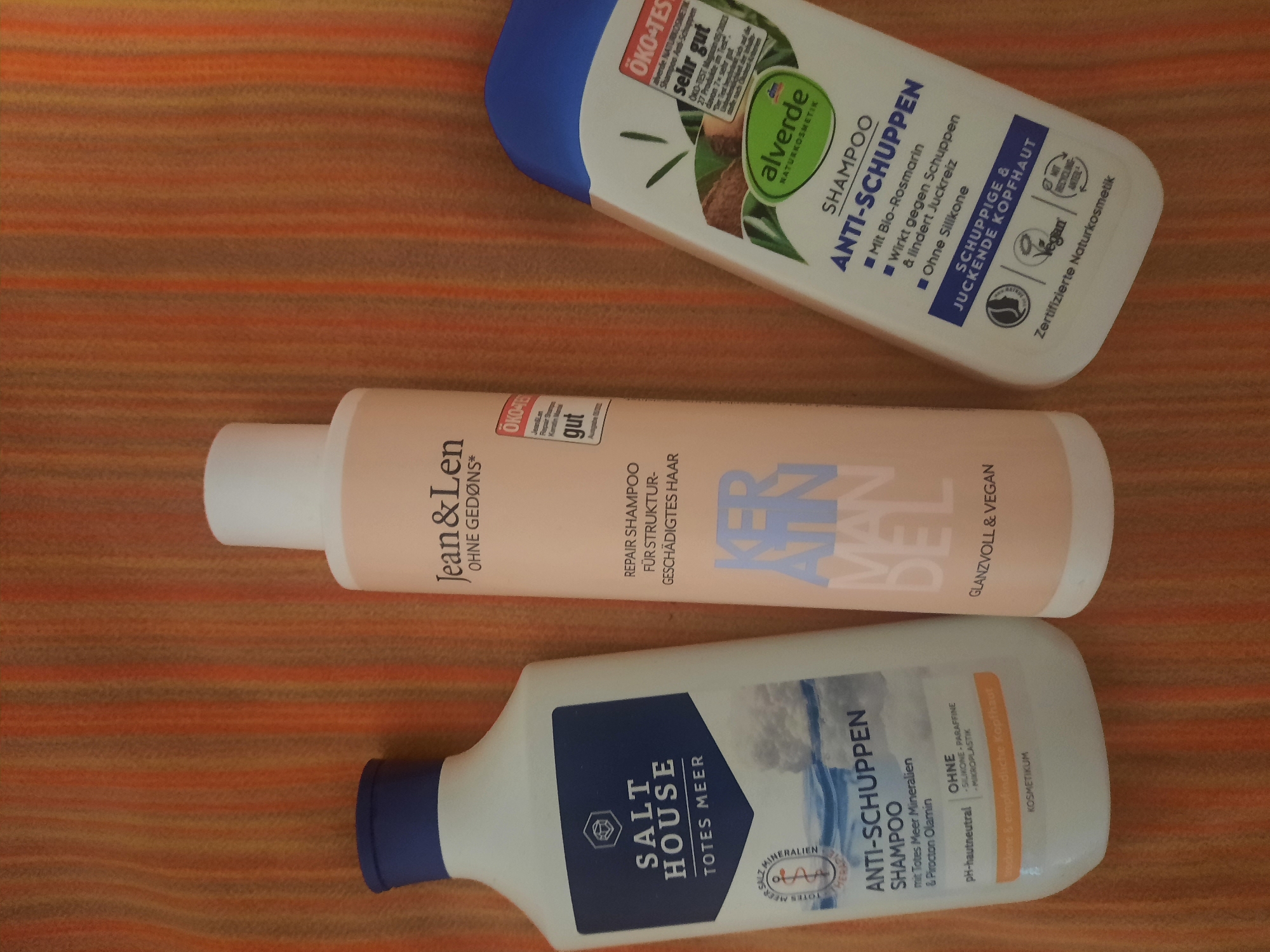Three bottles of shampoo are arranged on a textured, orange and brown striped surface. The bottle on the left is white with a dark blue label that reads "SALT HOUSE TOTES MEER" and "ANTI-SCHUPPEN SHAMPOO". The middle bottle is a pale pink with a white cap and a label that reads "Jean & Len" and "KERATIN MAN DEL". The bottle on the right is white with a blue top and a green and white label that reads "alverde NATURKOSMETIK" and "SHAMPOO ANTI-SCHUPPEN". The image does not contain any information about the city of Rosenheim, Germany.
